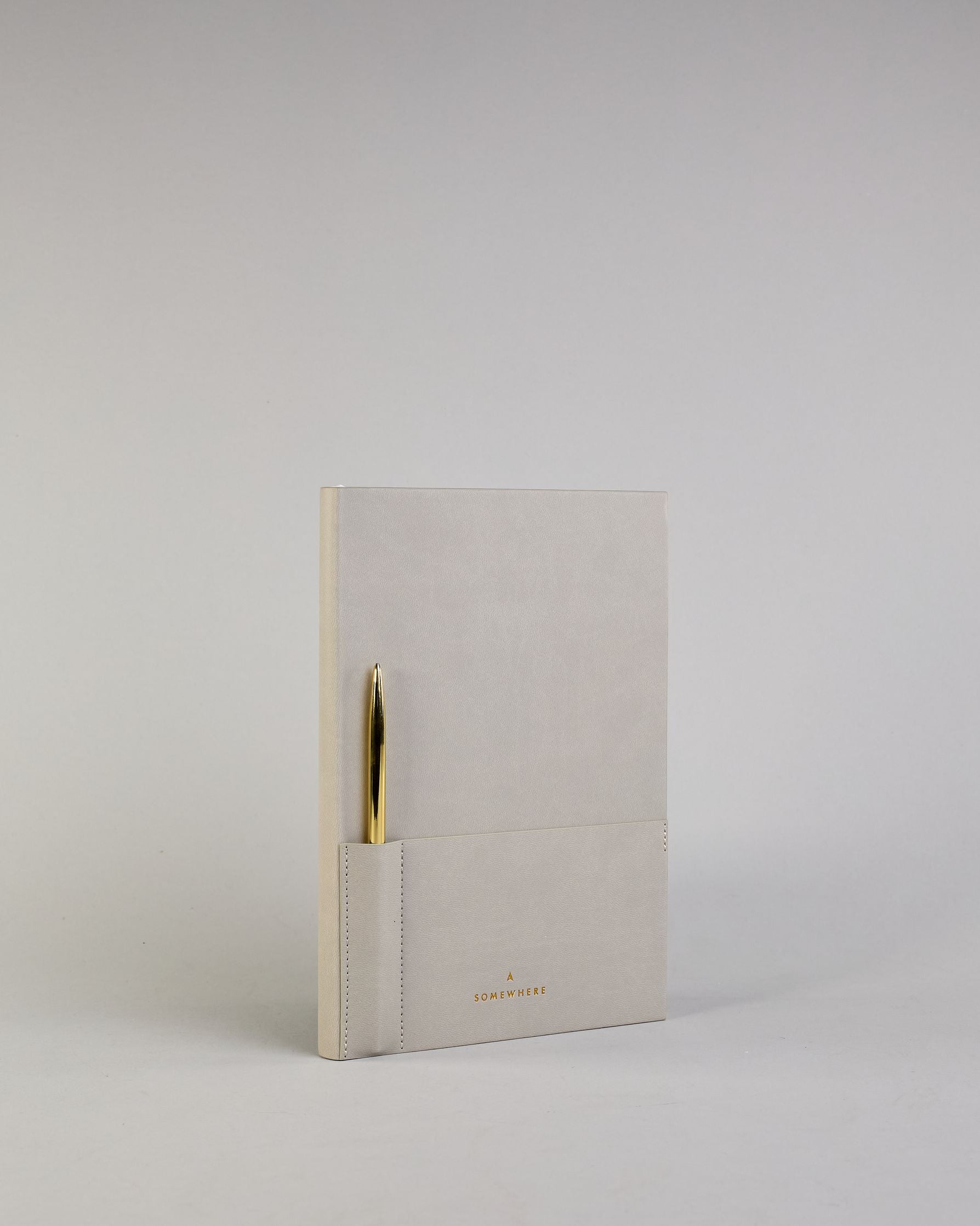 Hellgraues PU-Notizbuch mit zwei Laschen an der Vorderseite, eine Stiftschlaufe und einem zusätzlichem Fach, in dem auch ein Smartphone Platz findet. Unten dezent goldene Prägung mit dem Schriftzug ‚Somewhere‘. Ein goldfarbener Stift steckt seitlich in der Lasche. #17