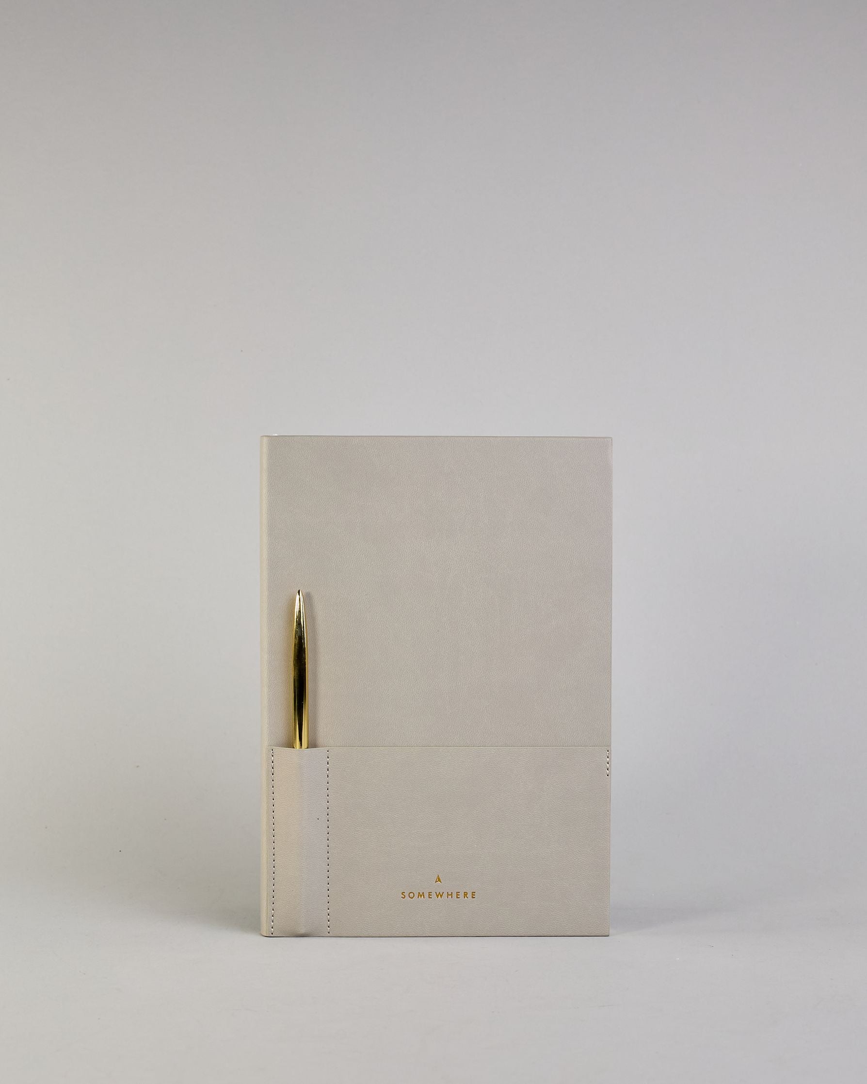 Hellgraues PU-Notizbuch mit zwei Laschen an der Vorderseite, eine Stiftschlaufe und einem zusätzlichem Fach, in dem auch ein Smartphone Platz findet. Unten dezent goldene Prägung mit dem Schriftzug ‚Somewhere‘. Ein goldfarbener Stift steckt seitlich in der Lasche. #16