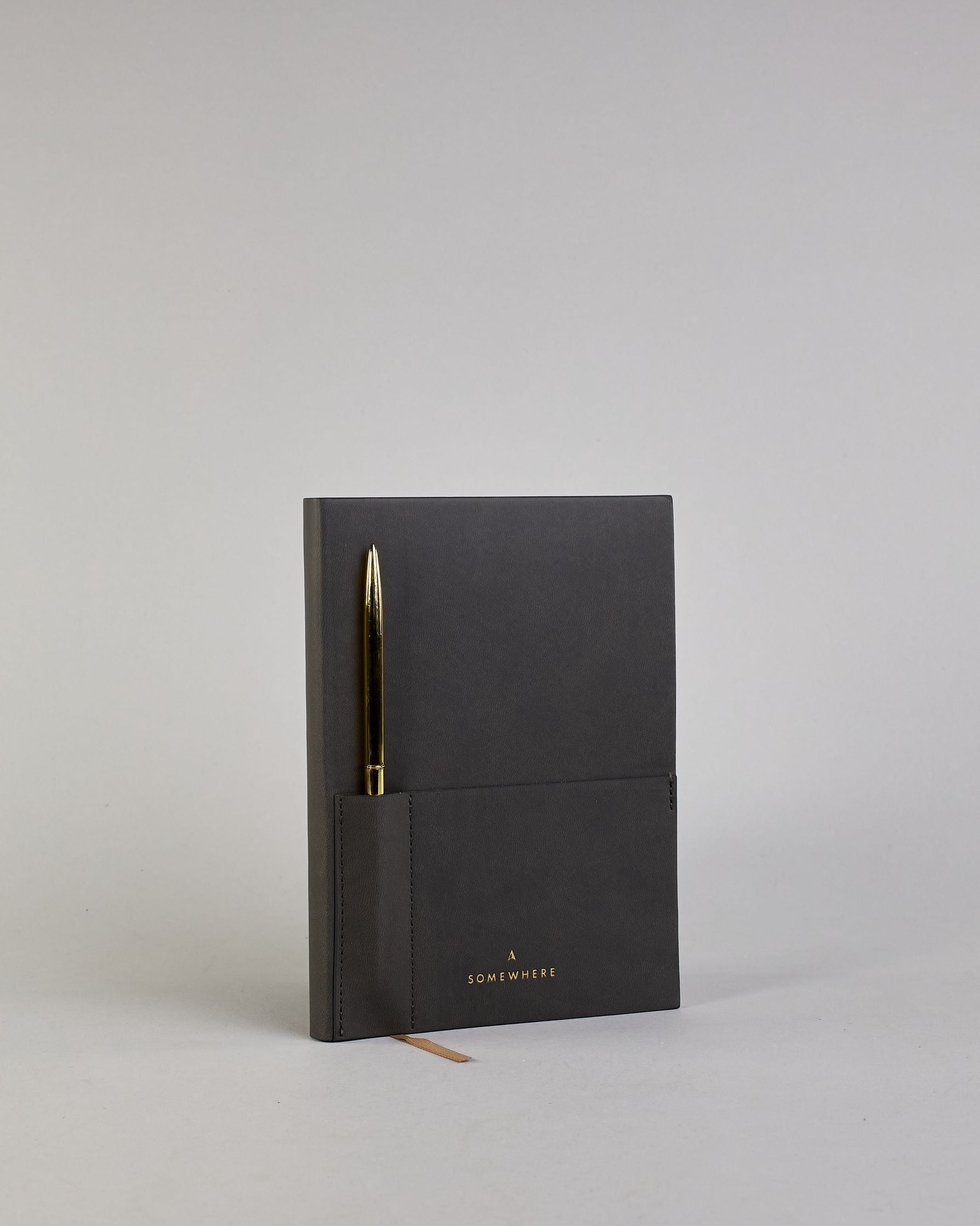 Schwarzes PU-Notizbuch mit zwei Laschen an der Vorderseite, eine Stiftschlaufe und einem zusätzlichem Fach, in dem auch ein Smartphone Platz findet. Unten dezent goldene Prägung mit dem Schriftzug ‚Somewhere‘. Ein goldfarbener Stift steckt seitlich in der Lasche. #2
