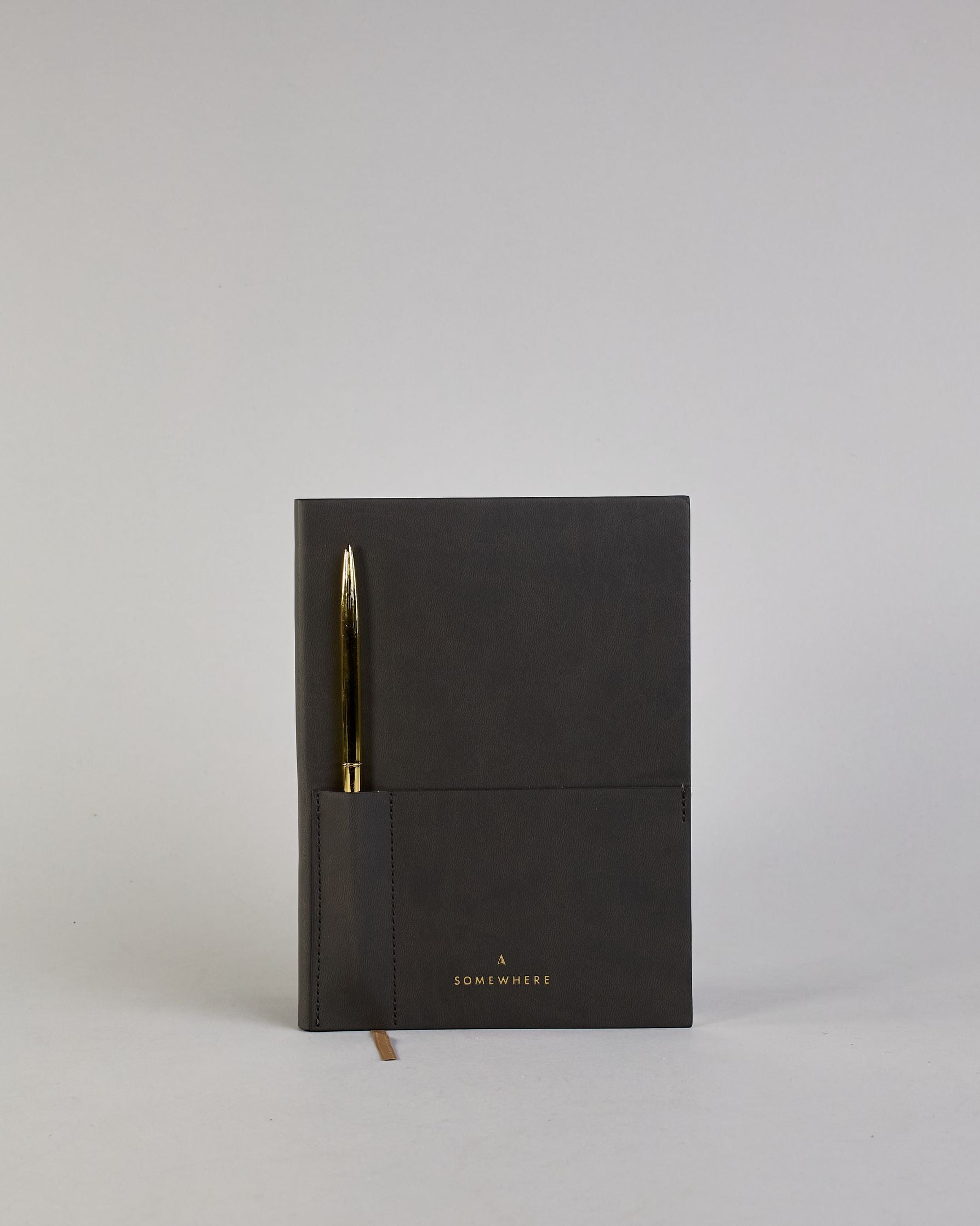 Schwarzes PU-Notizbuch mit zwei Laschen an der Vorderseite, eine Stiftschlaufe und einem zusätzlichem Fach, in dem auch ein Smartphone Platz findet. Unten dezent goldene Prägung mit dem Schriftzug ‚Somewhere‘. Ein goldfarbener Stift steckt seitlich in der Lasche. #1