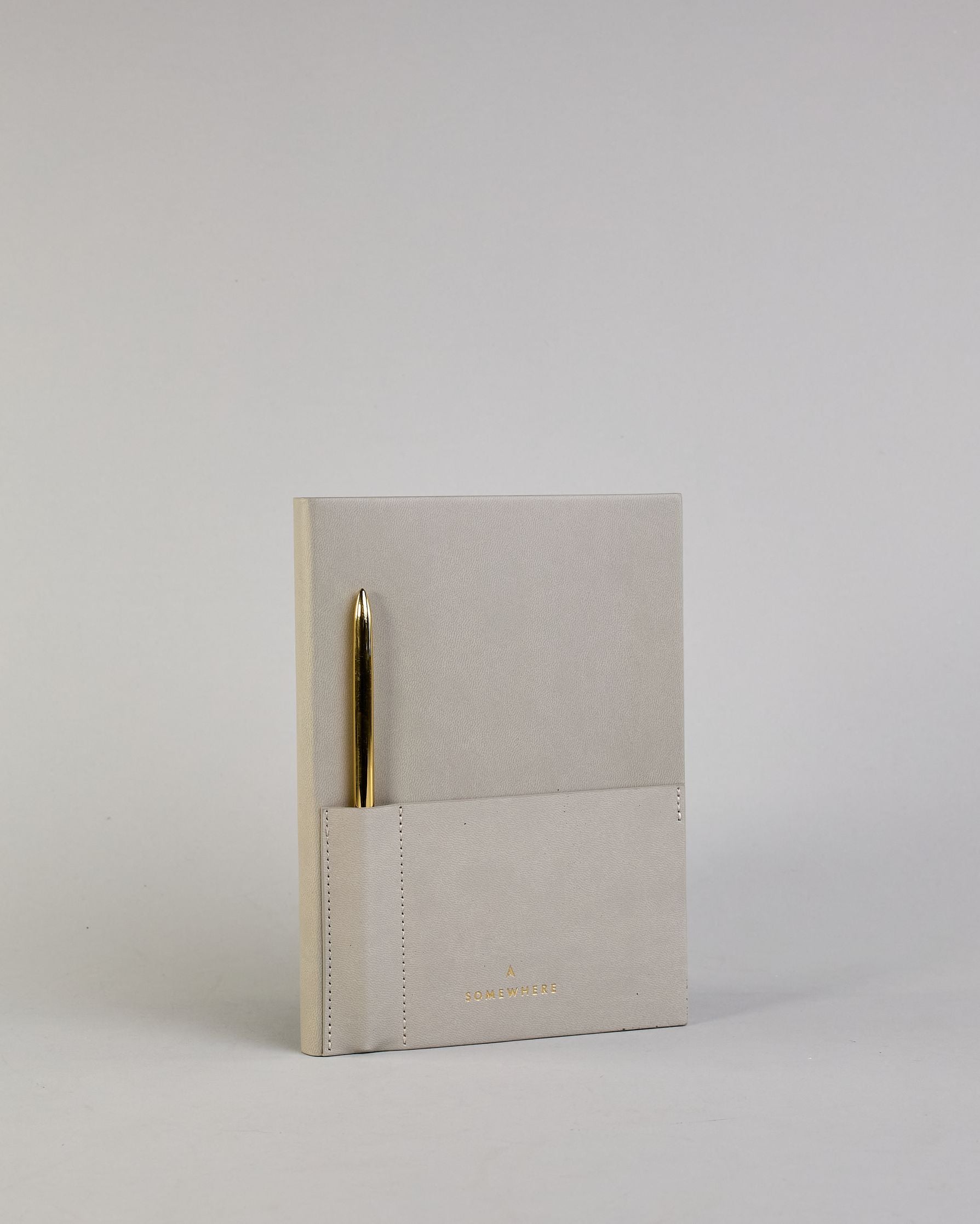 Hellgraues PU-Notizbuch mit zwei Laschen an der Vorderseite, eine Stiftschlaufe und einem zusätzlichem Fach, in dem auch ein Smartphone Platz findet. Unten dezent goldene Prägung mit dem Schriftzug ‚Somewhere‘. Ein goldfarbener Stift steckt seitlich in der Lasche. #8