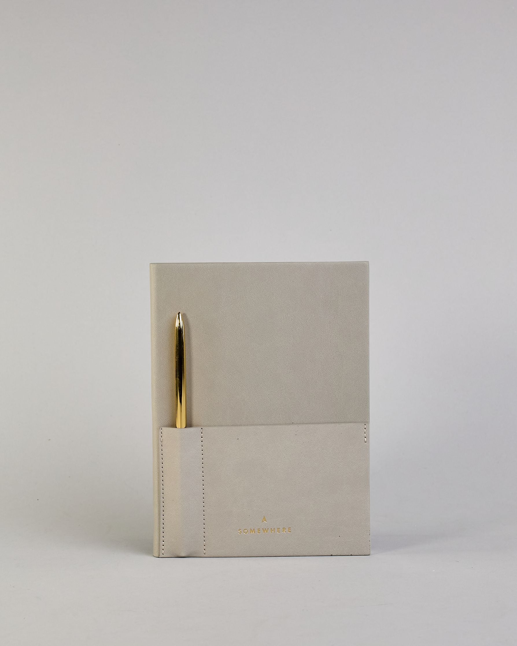 Hellgraues PU-Notizbuch mit zwei Laschen an der Vorderseite, eine Stiftschlaufe und einem zusätzlichem Fach, in dem auch ein Smartphone Platz findet. Unten dezent goldene Prägung mit dem Schriftzug ‚Somewhere‘. Ein goldfarbener Stift steckt seitlich in der Lasche. #7