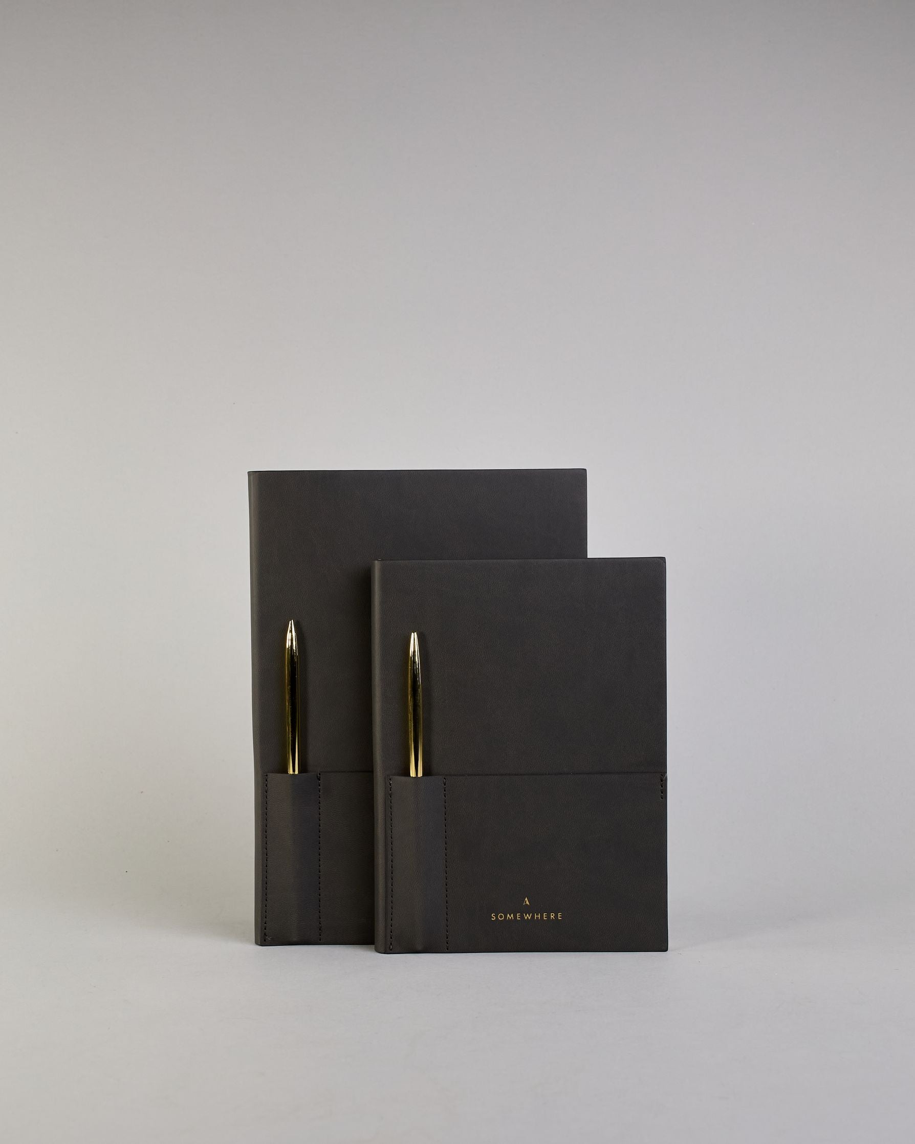Schwarzes PU-Notizbuch mit zwei Laschen an der Vorderseite, eine Stiftschlaufe und einem zusätzlichem Fach, in dem auch ein Smartphone Platz findet. Unten dezent goldene Prägung mit dem Schriftzug ‚Somewhere‘. Ein goldfarbener Stift steckt seitlich in der Lasche. #20
