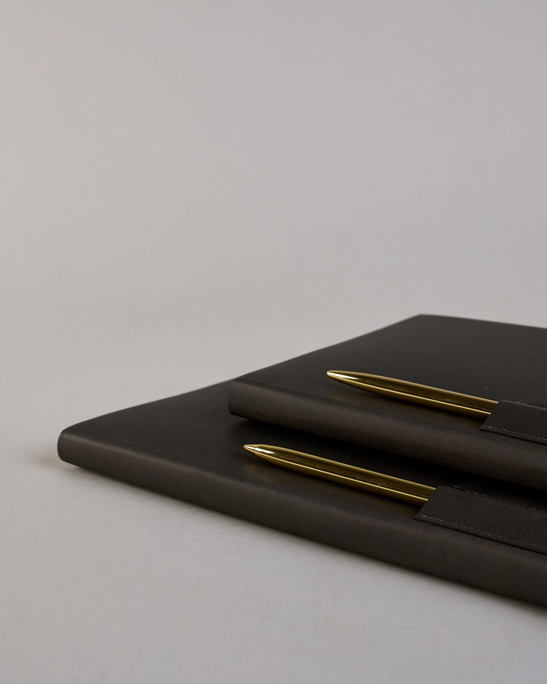 Schwarzes PU-Notizbuch mit zwei Laschen an der Vorderseite, eine Stiftschlaufe und einem zusätzlichem Fach, in dem auch ein Smartphone Platz findet. Unten dezent goldene Prägung mit dem Schriftzug ‚Somewhere‘. Ein goldfarbener Stift steckt seitlich in der Lasche. #21