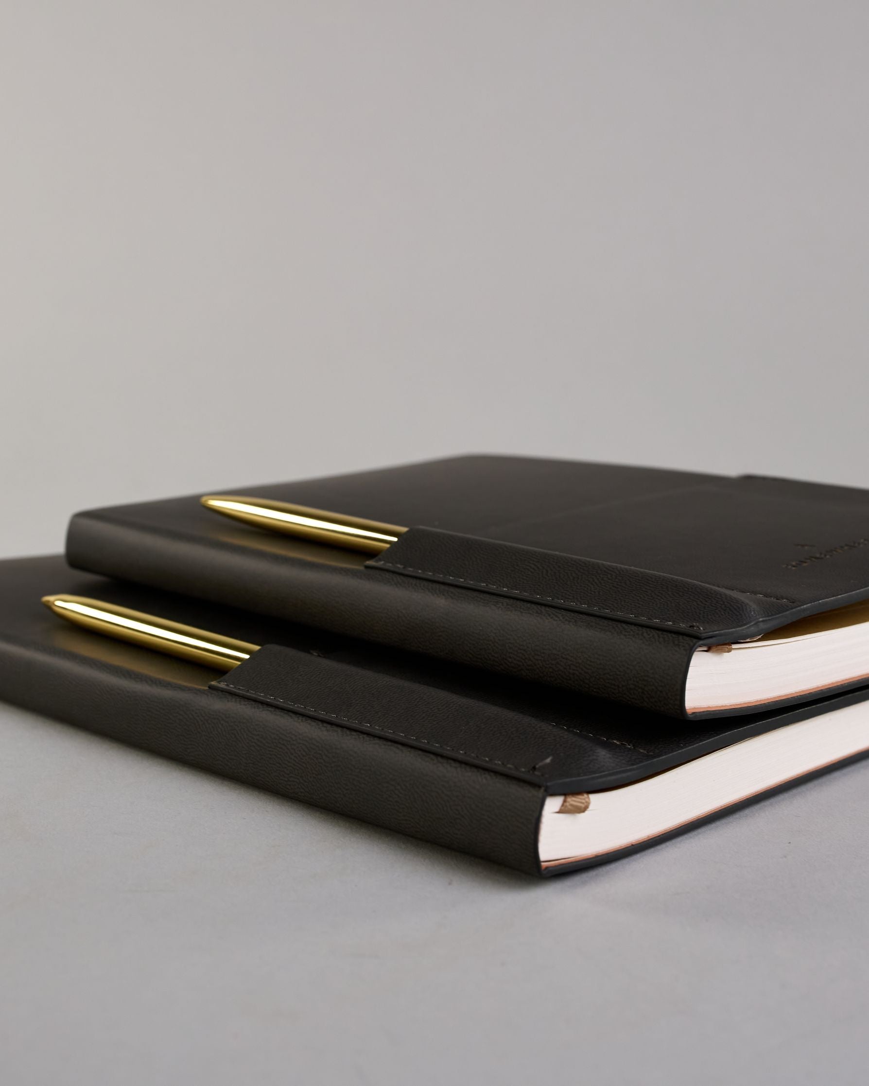 Schwarzes PU-Notizbuch mit zwei Laschen an der Vorderseite, eine Stiftschlaufe und einem zusätzlichem Fach, in dem auch ein Smartphone Platz findet. Unten dezent goldene Prägung mit dem Schriftzug ‚Somewhere‘. Ein goldfarbener Stift steckt seitlich in der Lasche. #22