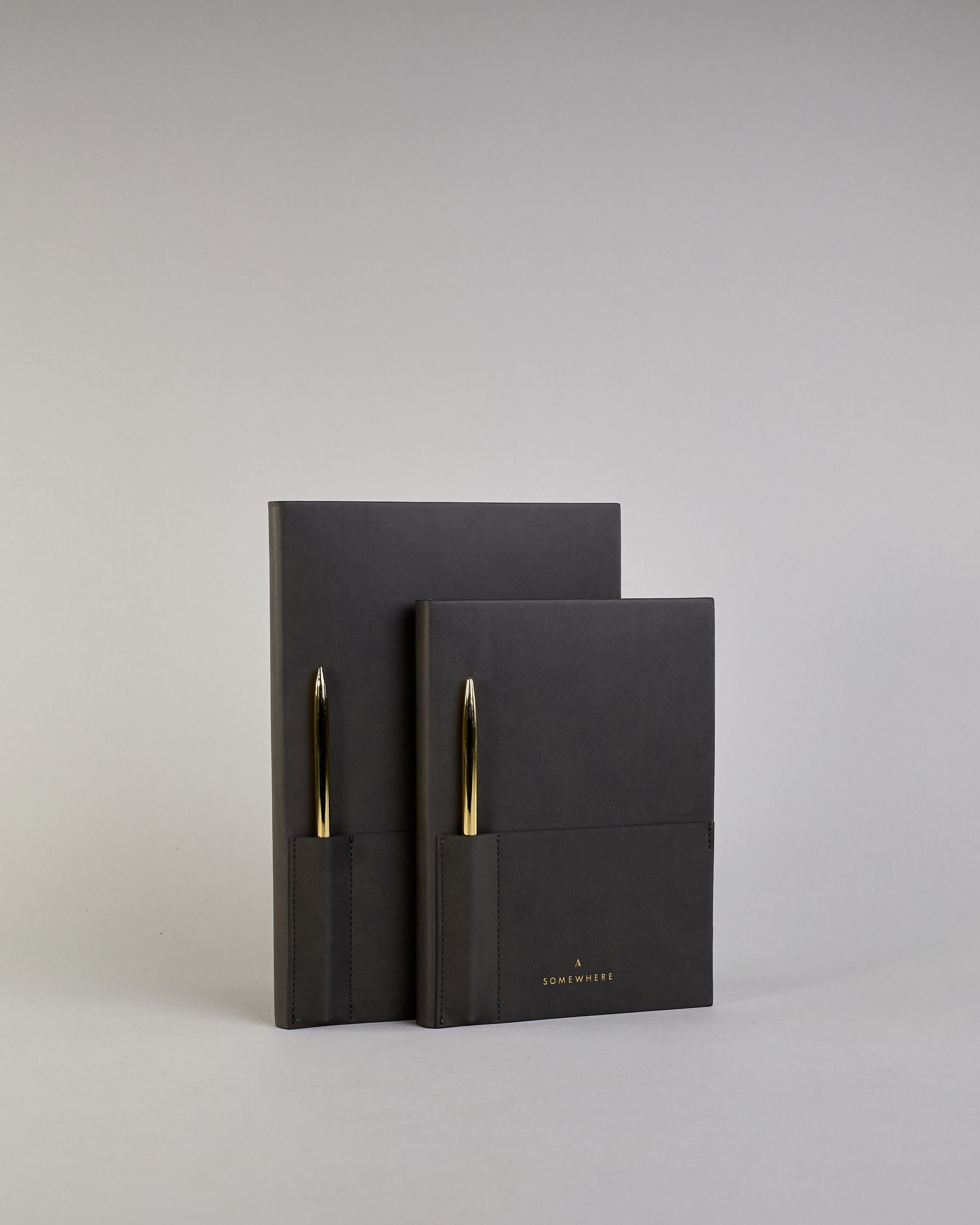 Schwarzes PU-Notizbuch mit zwei Laschen an der Vorderseite, eine Stiftschlaufe und einem zusätzlichem Fach, in dem auch ein Smartphone Platz findet. Unten dezent goldene Prägung mit dem Schriftzug ‚Somewhere‘. Ein goldfarbener Stift steckt seitlich in der Lasche. #19
