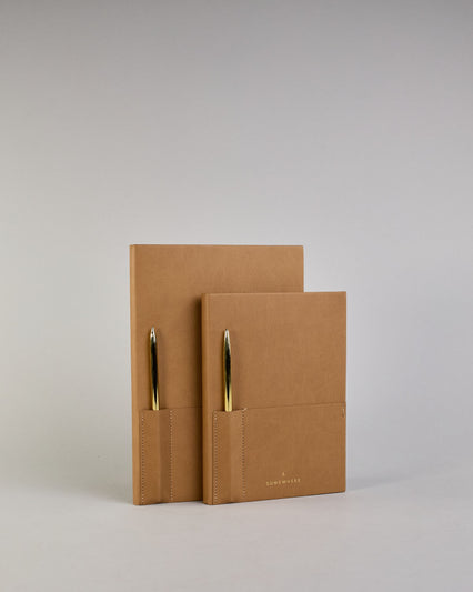 Cognacfarbenes PU-Notizbuch mit zwei Laschen an der Vorderseite, eine Stiftschlaufe und einem zusätzlichem Fach, in dem auch ein Smartphone Platz findet. Unten dezent goldene Prägung mit dem Schriftzug ‚Somewhere‘. Ein goldfarbener Stift steckt seitlich in der Lasche. #24