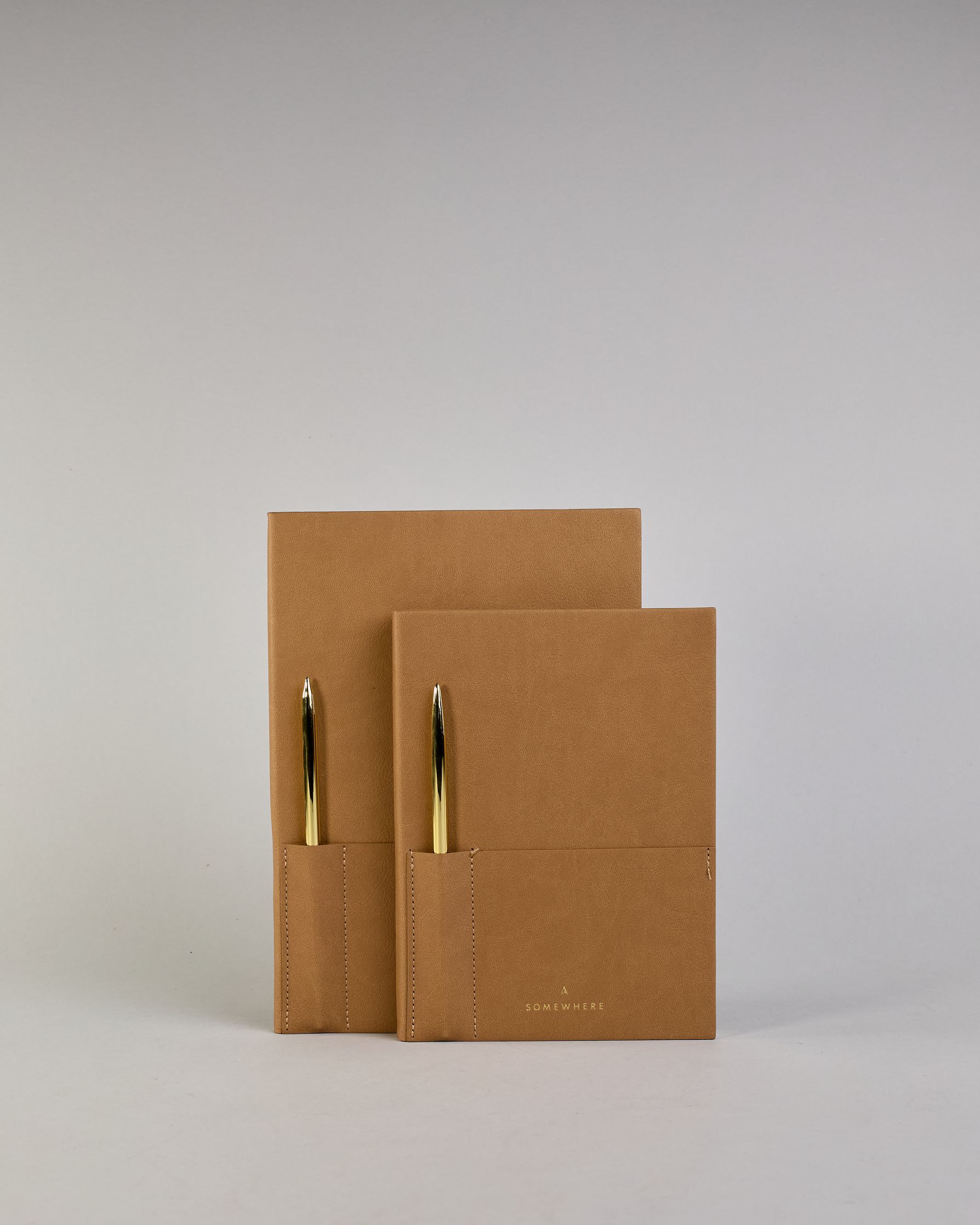 Cognacfarbenes PU-Notizbuch mit zwei Laschen an der Vorderseite, eine Stiftschlaufe und einem zusätzlichem Fach, in dem auch ein Smartphone Platz findet. Unten dezent goldene Prägung mit dem Schriftzug ‚Somewhere‘. Ein goldfarbener Stift steckt seitlich in der Lasche. #25