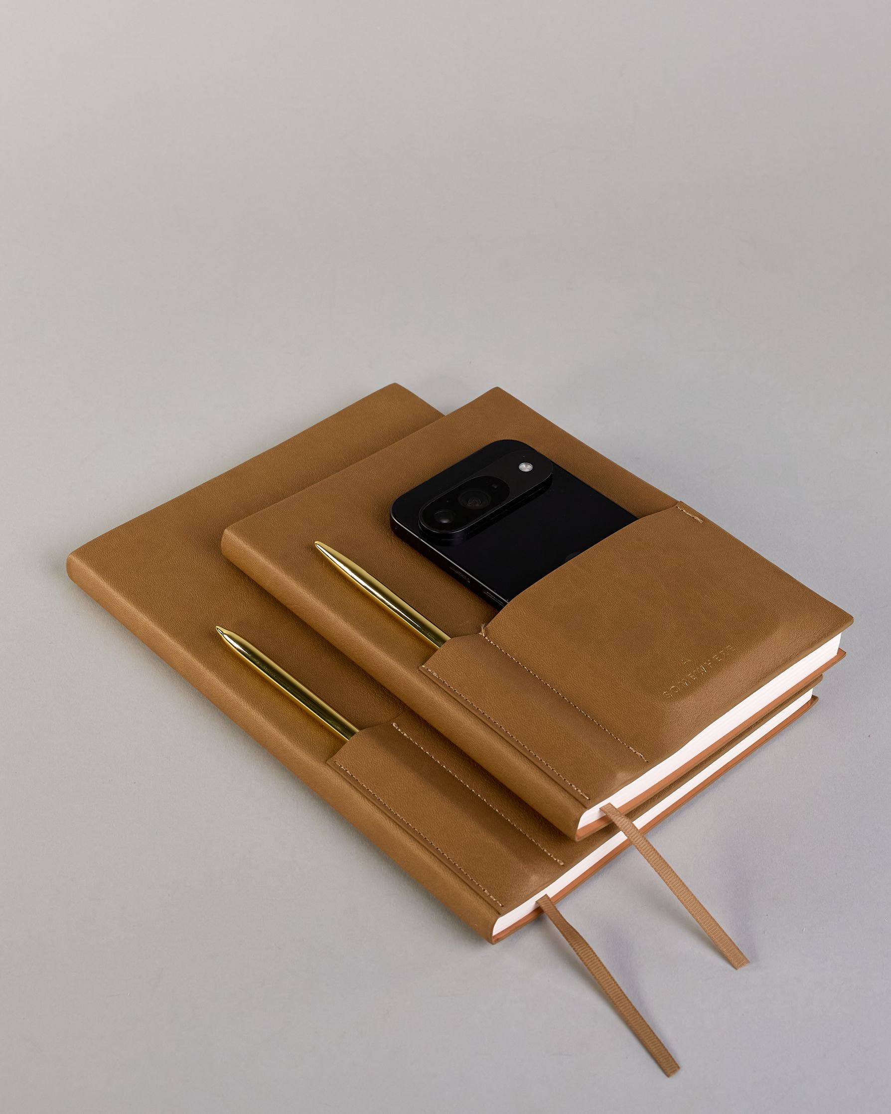 Cognacfarbenes PU-Notizbuch mit zwei Laschen an der Vorderseite, eine Stiftschlaufe und einem zusätzlichem Fach, in dem auch ein Smartphone Platz findet. Unten dezent goldene Prägung mit dem Schriftzug ‚Somewhere‘. Ein goldfarbener Stift steckt seitlich in der Lasche. #26