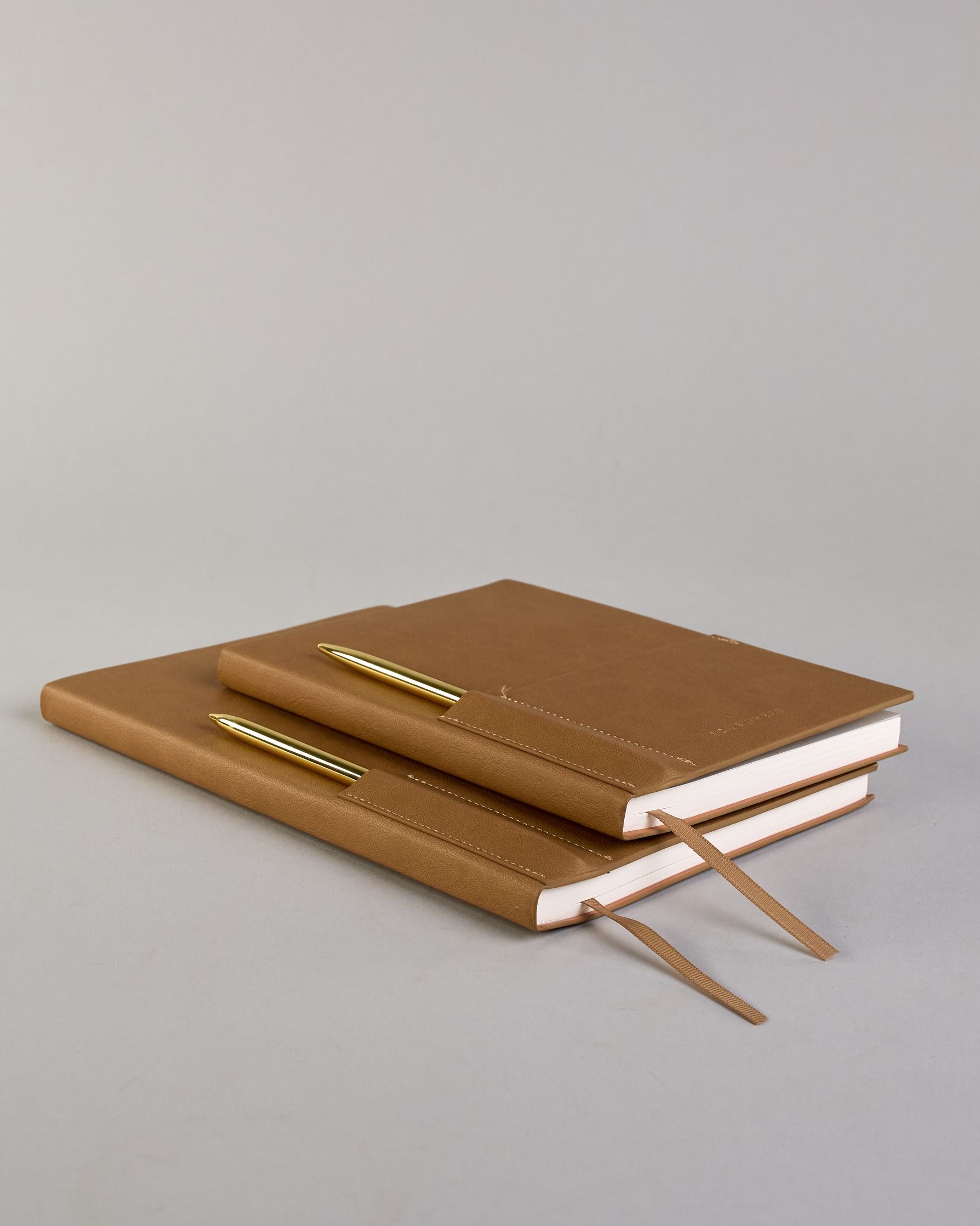 Cognacfarbenes PU-Notizbuch mit zwei Laschen an der Vorderseite, eine Stiftschlaufe und einem zusätzlichem Fach, in dem auch ein Smartphone Platz findet. Unten dezent goldene Prägung mit dem Schriftzug ‚Somewhere‘. Ein goldfarbener Stift steckt seitlich in der Lasche. #27