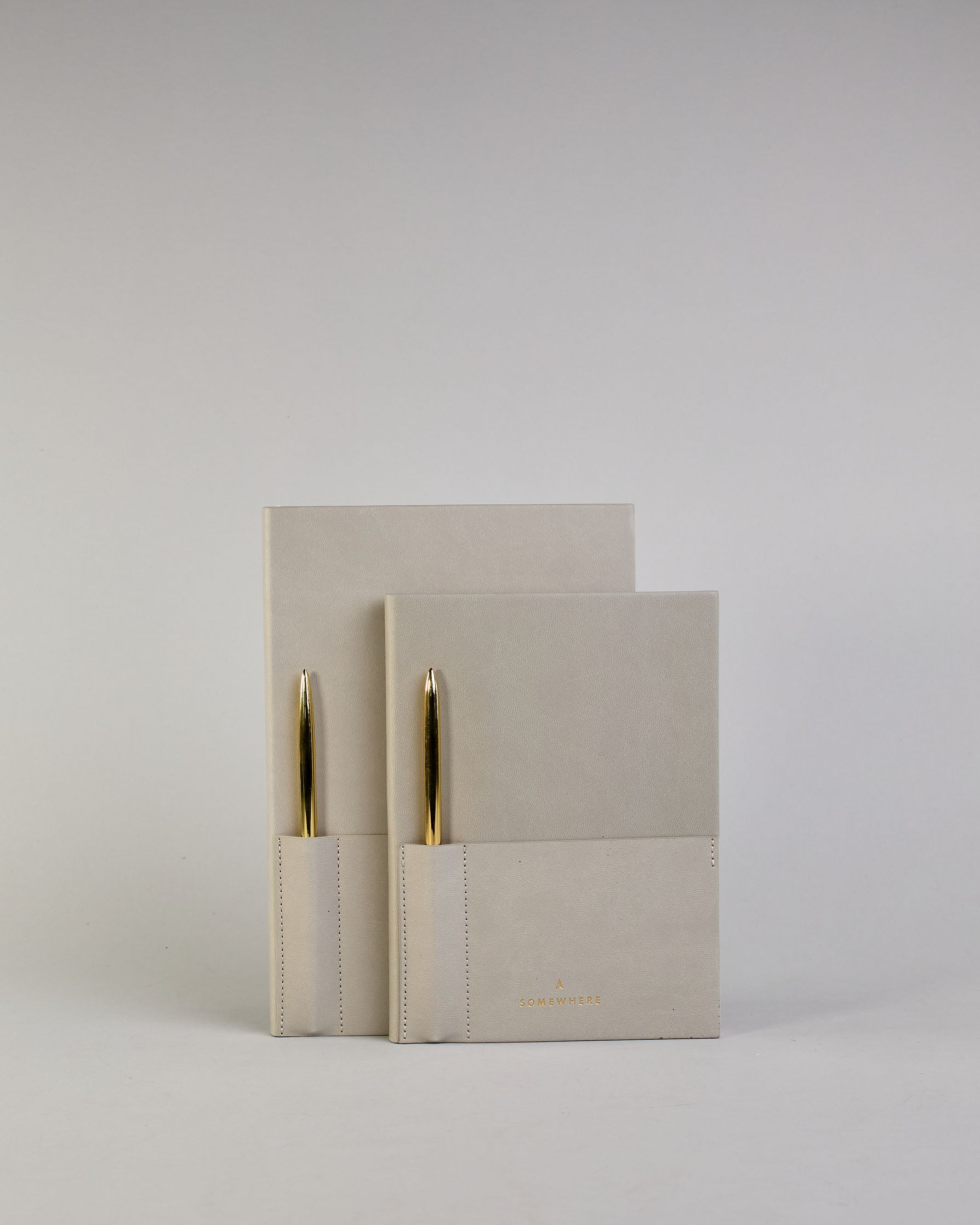 Hellgraues PU-Notizbuch mit zwei Laschen an der Vorderseite, eine Stiftschlaufe und einem zusätzlichem Fach, in dem auch ein Smartphone Platz findet. Unten dezent goldene Prägung mit dem Schriftzug ‚Somewhere‘. Ein goldfarbener Stift steckt seitlich in der Lasche. #30