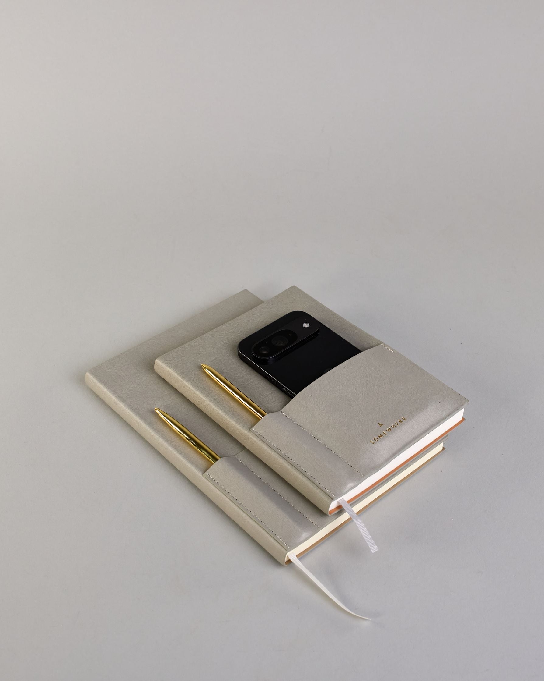 Hellgraues PU-Notizbuch mit zwei Laschen an der Vorderseite, eine Stiftschlaufe und einem zusätzlichem Fach, in dem auch ein Smartphone Platz findet. Unten dezent goldene Prägung mit dem Schriftzug ‚Somewhere‘. Ein goldfarbener Stift steckt seitlich in der Lasche. #31
