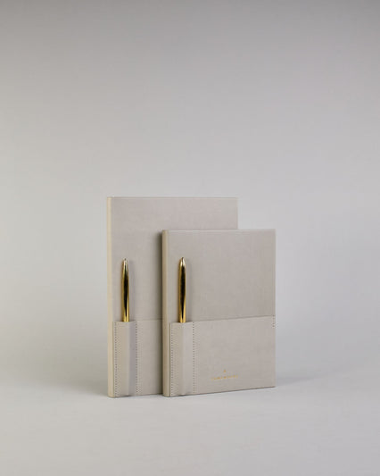 Hellgraues PU-Notizbuch mit zwei Laschen an der Vorderseite, eine Stiftschlaufe und einem zusätzlichem Fach, in dem auch ein Smartphone Platz findet. Unten dezent goldene Prägung mit dem Schriftzug ‚Somewhere‘. Ein goldfarbener Stift steckt seitlich in der Lasche. #29