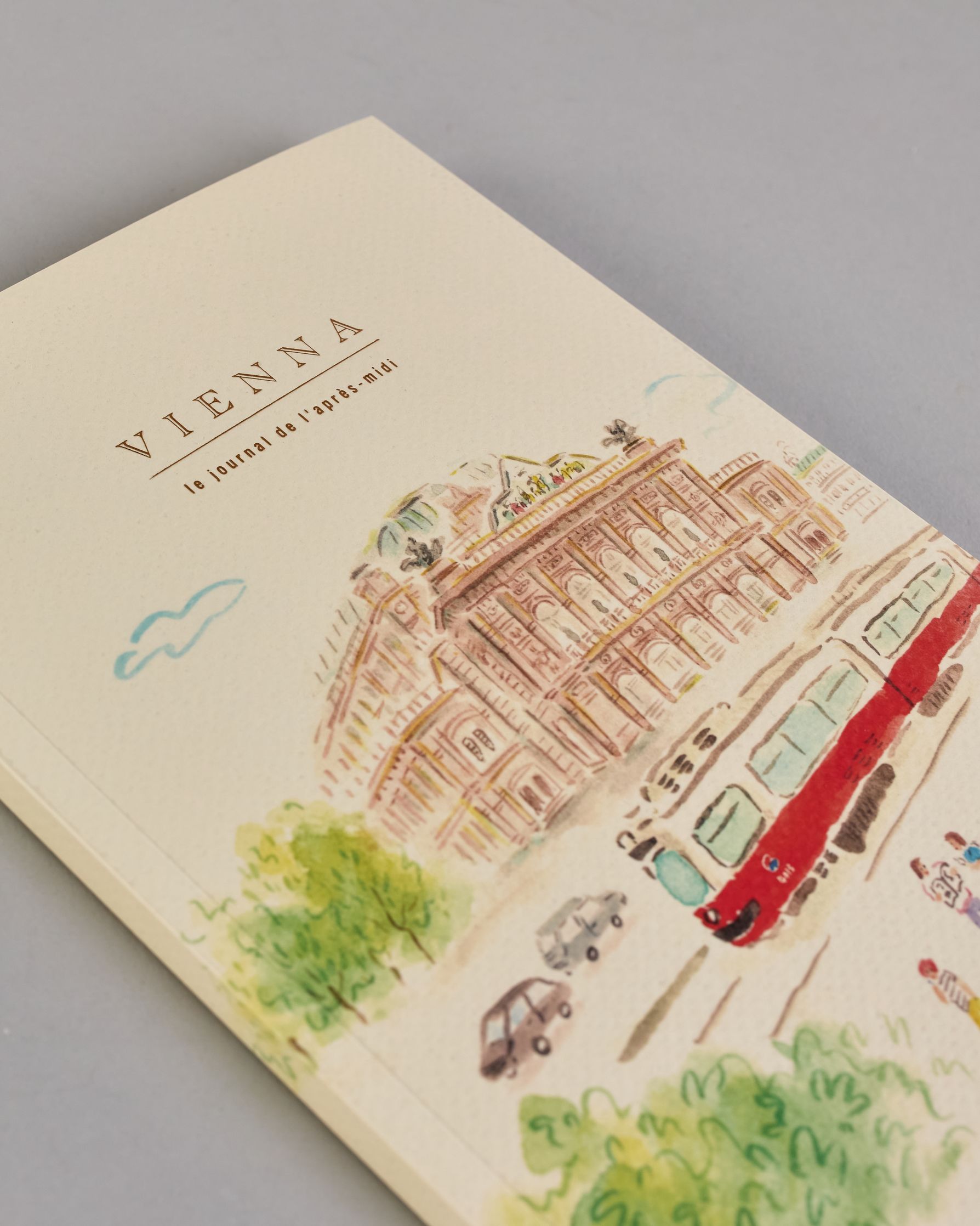 Notizbuch mit cremefarbenem Einband, darauf eine zarte Illustration der Wiener Staatsoper mit vorbeifahrender Straßenbahn und kleinen Figuren im Vordergrund. Titel: ‚Vienna – le journal de l’après-midi. Innen ist jede Seite individuell illustriert. #3