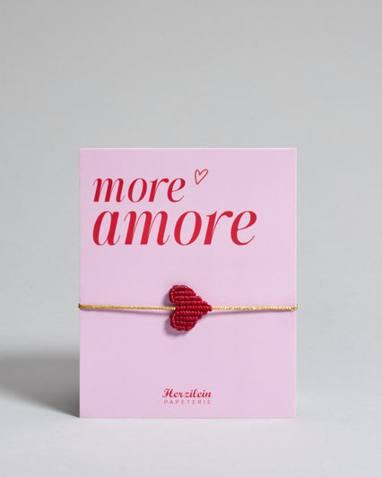Armband mit goldenem Band und rotem Makramee geflochtenem Herz. Das Armband ist auf einer Karte auf der "more amore" steht. Die Karte ist pink und rot gehalten. 