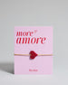 Armband mit goldenem Band und rotem Makramee geflochtenem Herz. Das Armband ist auf einer Karte auf der "more amore" steht. Die Karte ist pink und rot gehalten. 