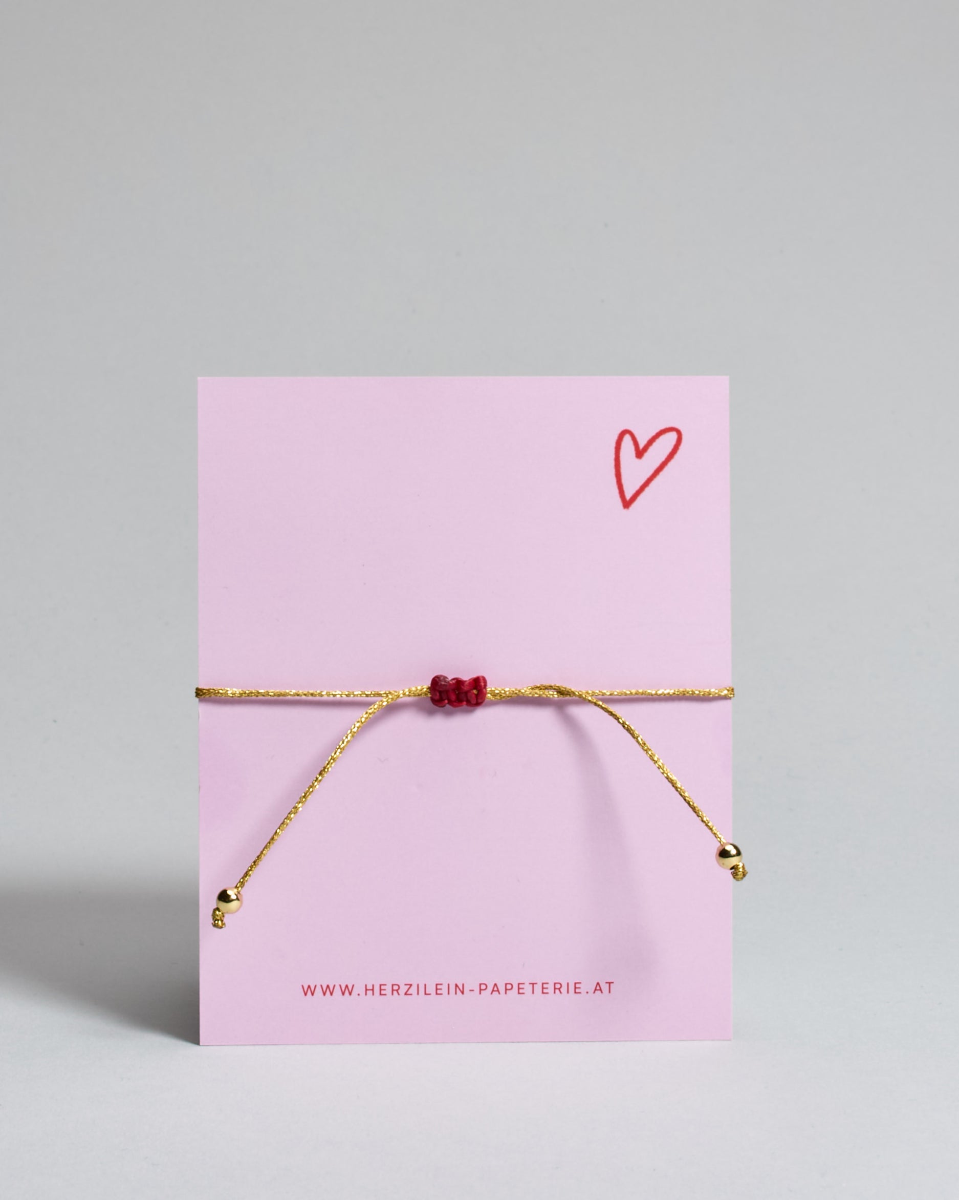 Armband mit goldenem Band und rotem Makramee geflochtenem Herz. Das Armband ist auf einer Karte auf der "more amore" steht. Die Karte ist pink und rot gehalten. 