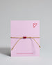 Armband mit goldenem Band und rotem Makramee geflochtenem Herz. Das Armband ist auf einer Karte auf der "more amore" steht. Die Karte ist pink und rot gehalten. 