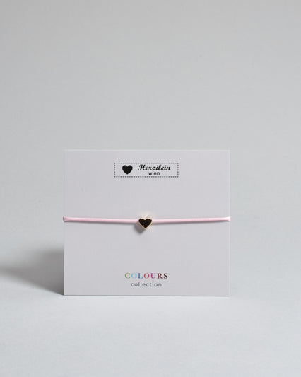 Armband mit rosa gewachstem Baumwollband und kleinem goldenem Herz Anhänger. Das Armband ist auf einer grauen Karte auf der "Herzilein-Wien, Colours-Collection" steht. 