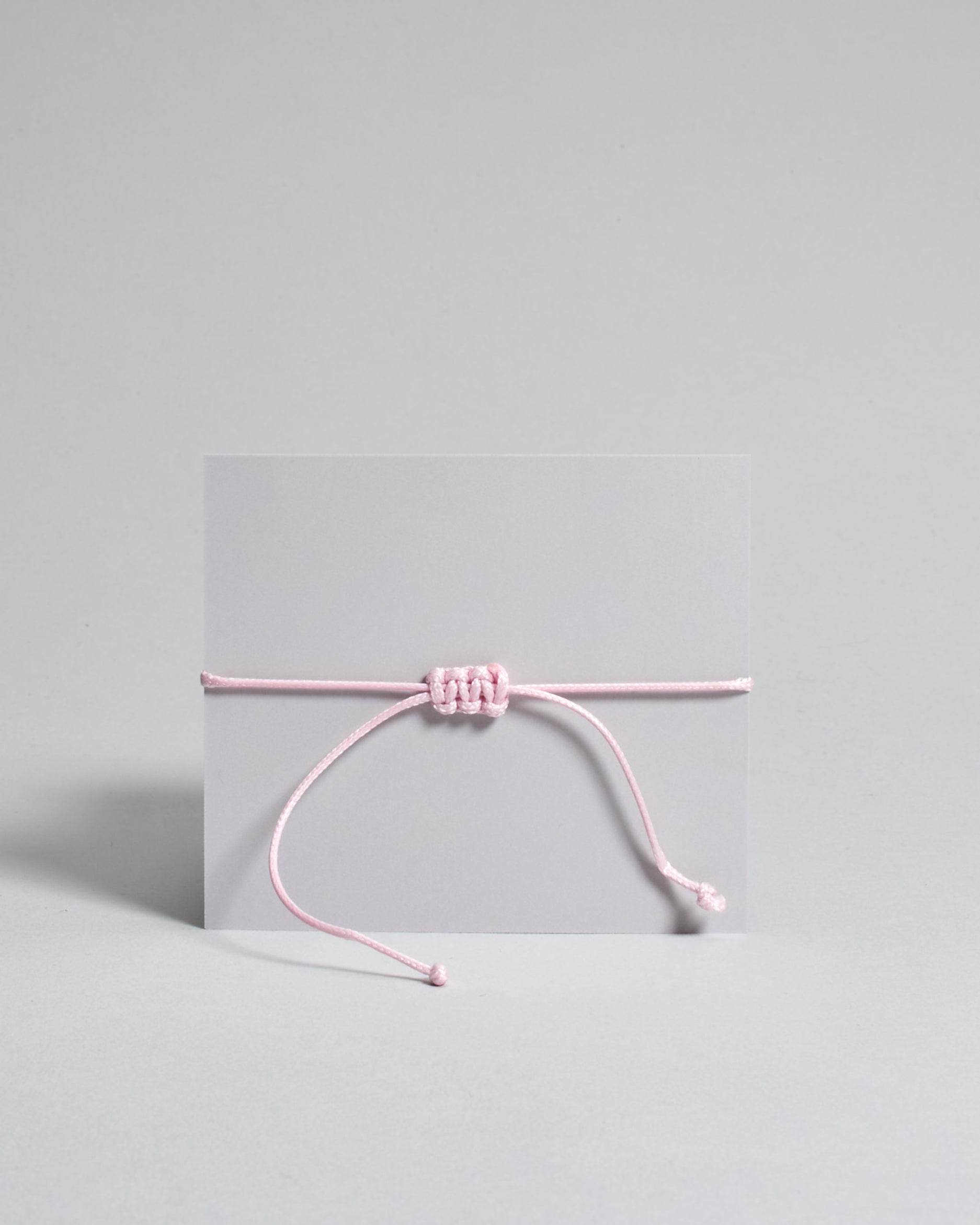 Armband mit rosa gewachstem Baumwollband und kleinem goldenem Herz Anhänger. Das Armband ist auf einer grauen Karte auf der "Herzilein-Wien, Colours-Collection" steht. 