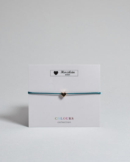 Armband mit petrol farbenem  gewachstem Baumwollband und kleinem goldenem Herz Anhänger. Das Armband ist auf einer grauen Karte auf der "Herzilein-Wien, Colours-Collection" steht. 