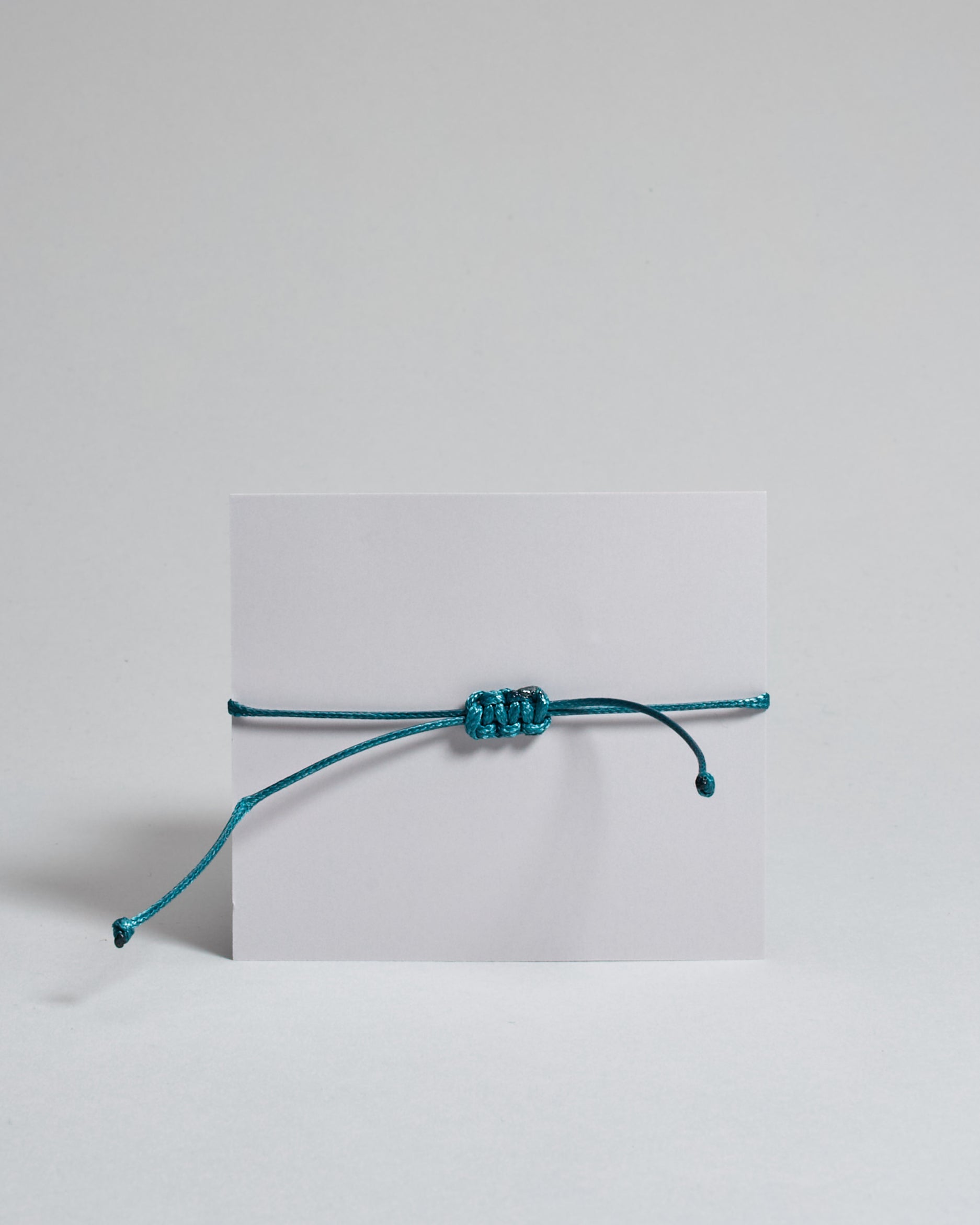Armband mit petrol farbenem  gewachstem Baumwollband und kleinem goldenem Herz Anhänger. Das Armband ist auf einer grauen Karte auf der "Herzilein-Wien, Colours-Collection" steht. 