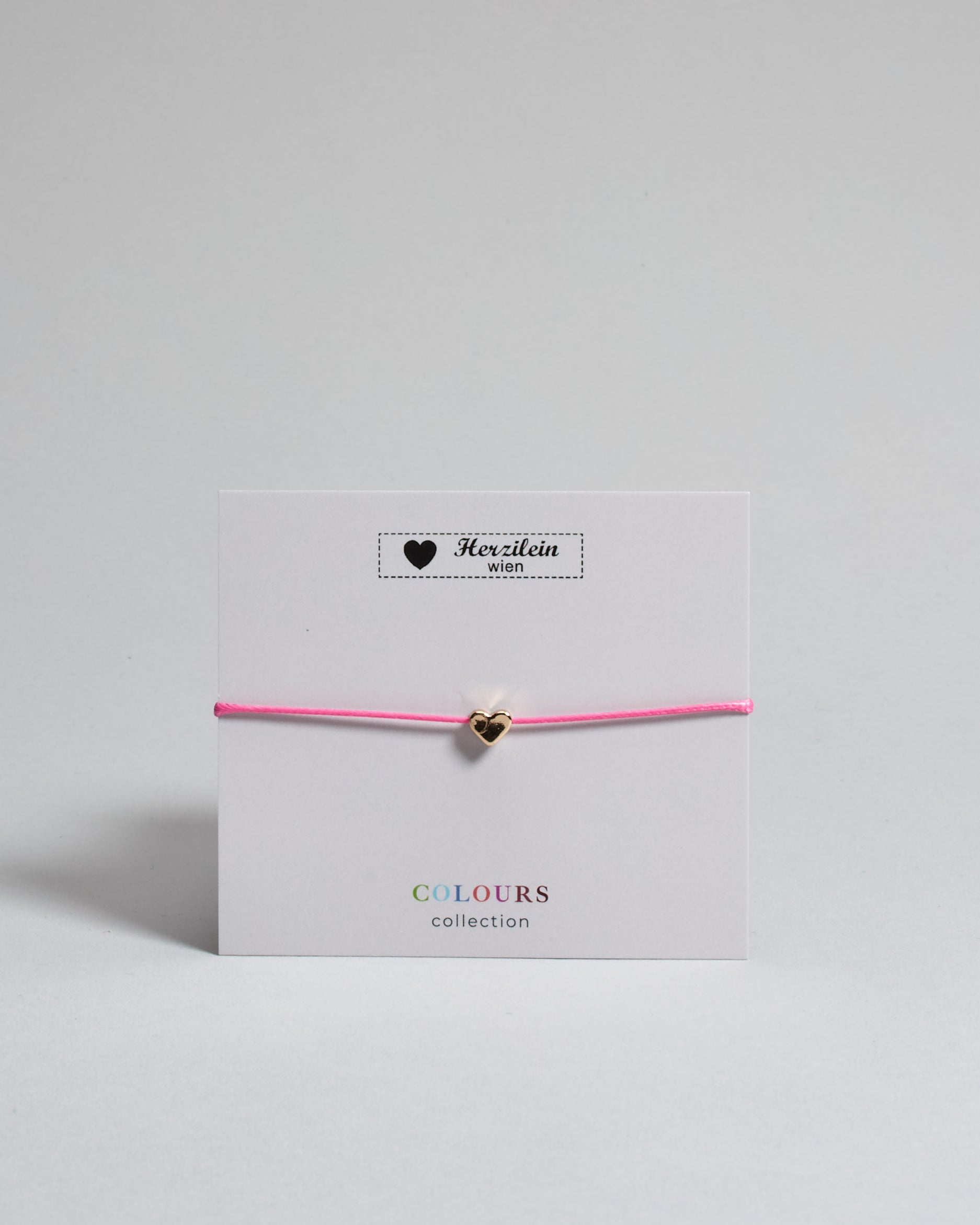 Armband mit pinkem gewachstem Baumwollband und kleinem goldenem Herz Anhänger. Das Armband ist auf einer grauen Karte auf der "Herzilein-Wien, Colours-Collection" steht. 