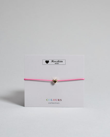 Armband mit pinkem gewachstem Baumwollband und kleinem goldenem Herz Anhänger. Das Armband ist auf einer grauen Karte auf der "Herzilein-Wien, Colours-Collection" steht. 