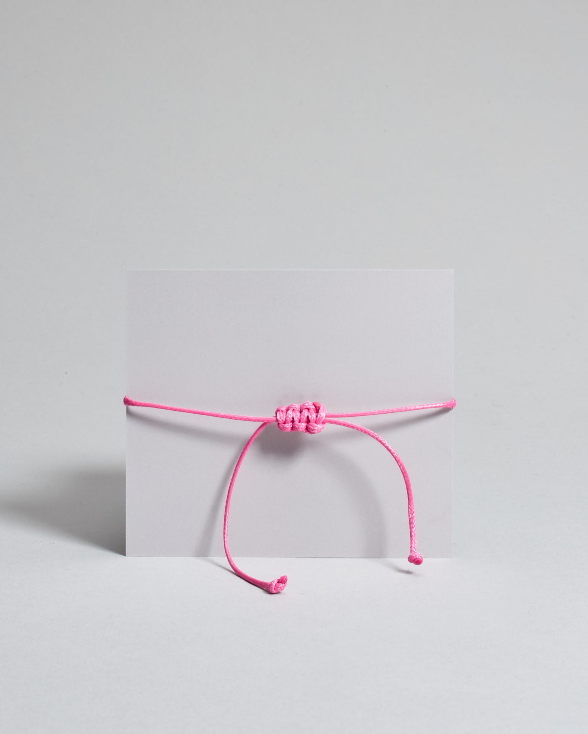 Armband mit pinkem gewachstem Baumwollband und kleinem goldenem Herz Anhänger. Das Armband ist auf einer grauen Karte auf der "Herzilein-Wien, Colours-Collection" steht. 