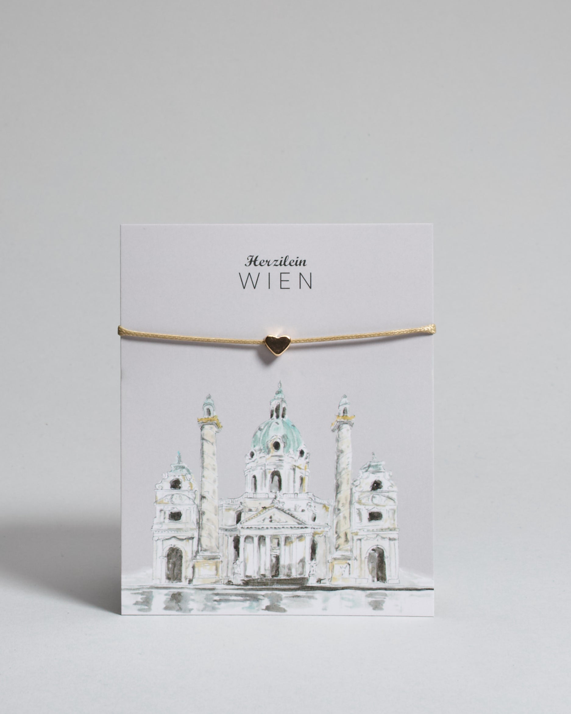 Armband mit beigem gewachstem Baumwollband und kleinem goldenem Herz Anhänger. Das Armband ist auf einer grauen Karte auf der "Herzilein-Wien" steht und unten die Karlskirche abgebildet ist. 