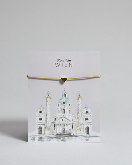 Armband mit beigem gewachstem Baumwollband und kleinem goldenem Herz Anhänger. Das Armband ist auf einer grauen Karte auf der "Herzilein-Wien" steht und unten die Karlskirche abgebildet ist. 