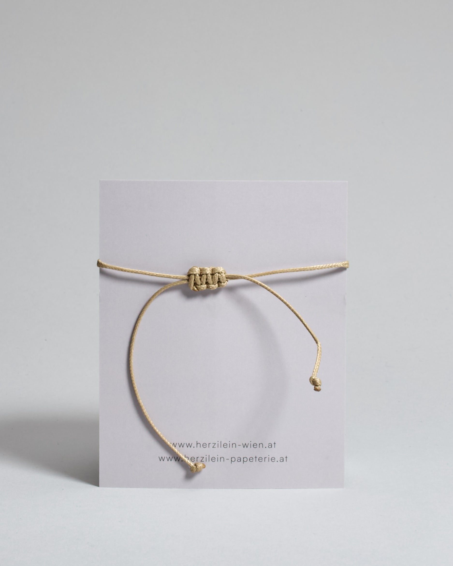 Armband mit beigem gewachstem Baumwollband und kleinem goldenem Herz Anhänger. Das Armband ist auf einer grauen Karte auf der "Herzilein-Wien" steht und unten die Karlskirche abgebildet ist. 