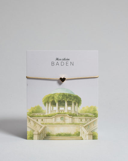 Armband mit beigem gewachstem Baumwollband und kleinem goldenem Herz Anhänger. Das Armband ist auf einer grauen Karte auf der "Herzilein-Wien" steht und unten die Beethofentempel abgebildet ist. 