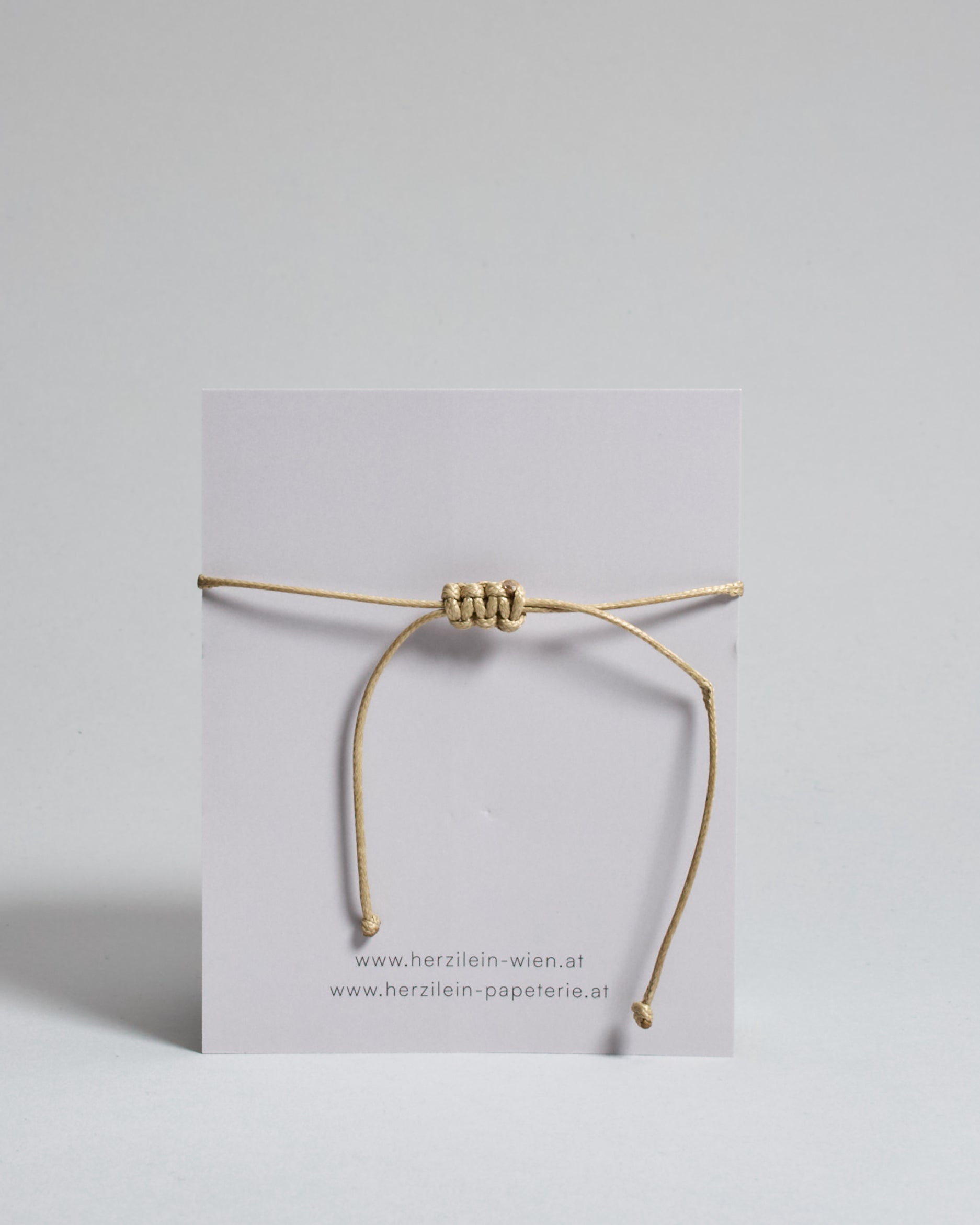 Armband mit beigem gewachstem Baumwollband und kleinem goldenem Herz Anhänger. Das Armband ist auf einer grauen Karte auf der "Herzilein-Wien" steht und unten die Beethofentempel abgebildet ist. 