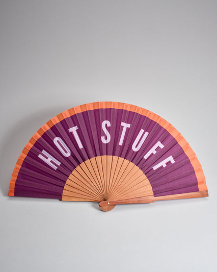 Fächer aus geöltem Holz mit Stoffbezug der oben einen orangen Rand hat und sonst lila ist. Es steht große "Hot Stuff" drauf. 
