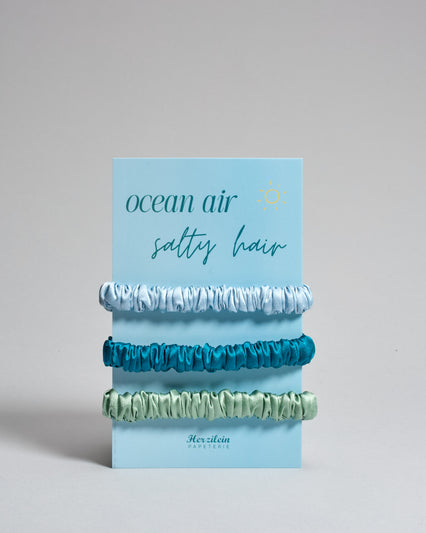 Drei Haar-Scrunchies aus Seide auf einer Karte auf der oben "ocean air, salty hair" steht. Die Scrunchies sind hellbau, petrol und mintfarben. 