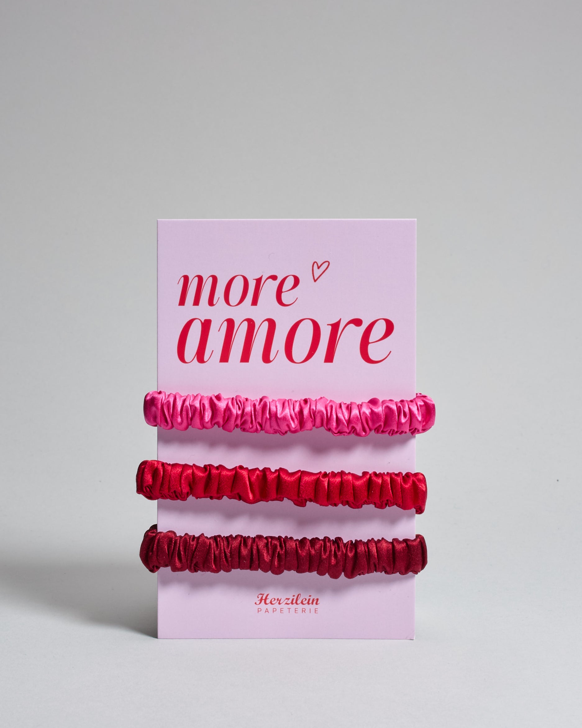 Drei Haar-Scrunchies aus Seide auf einer Karte auf der oben "more amore" steht. Die Scrunchies sind pink, rot und bordeaux. 