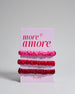 Drei Haar-Scrunchies aus Seide auf einer Karte auf der oben "more amore" steht. Die Scrunchies sind pink, rot und bordeaux. 