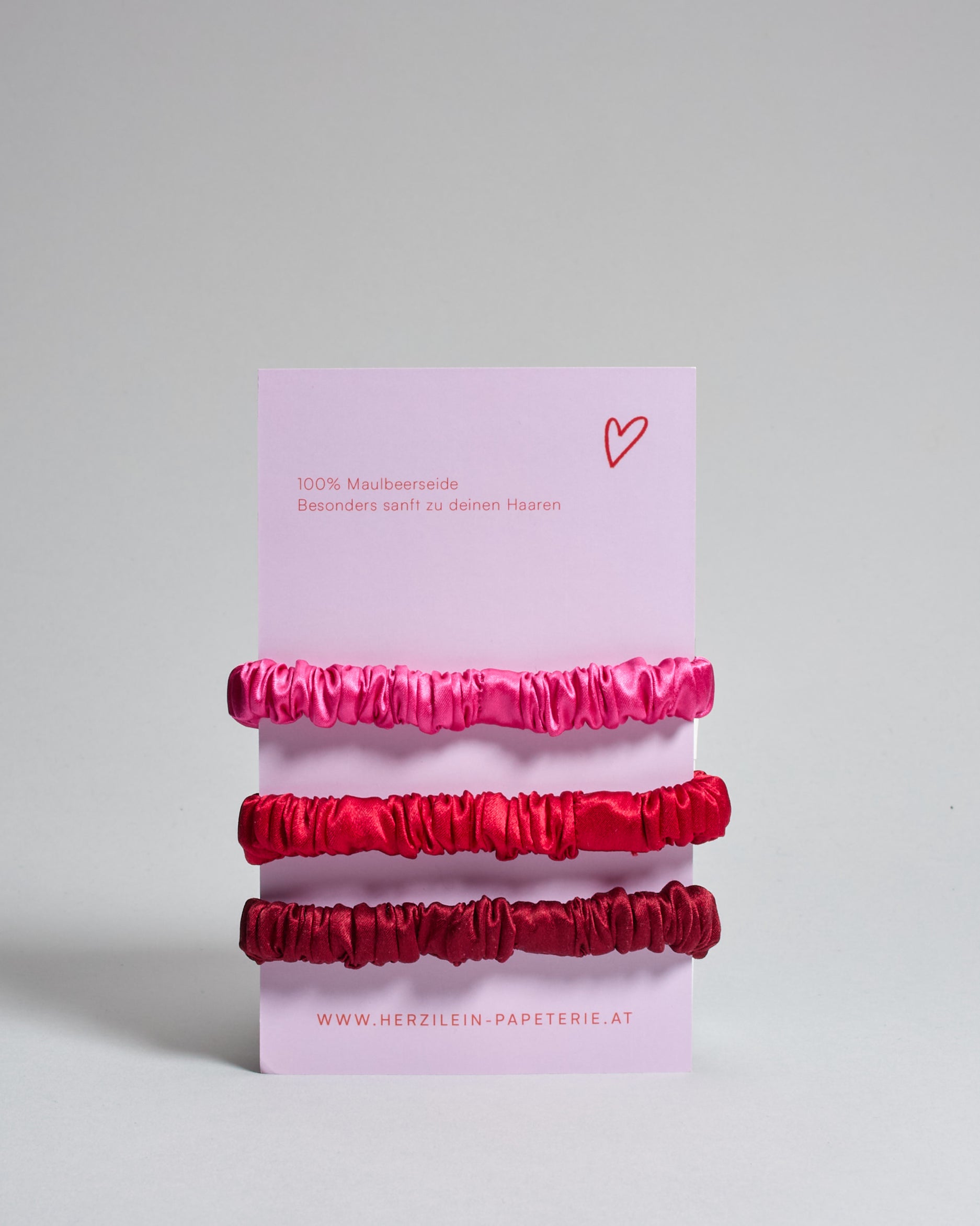 Drei Haar-Scrunchies aus Seide auf einer Karte auf der oben "more amore" steht. Die Scrunchies sind pink, rot und bordeaux. 