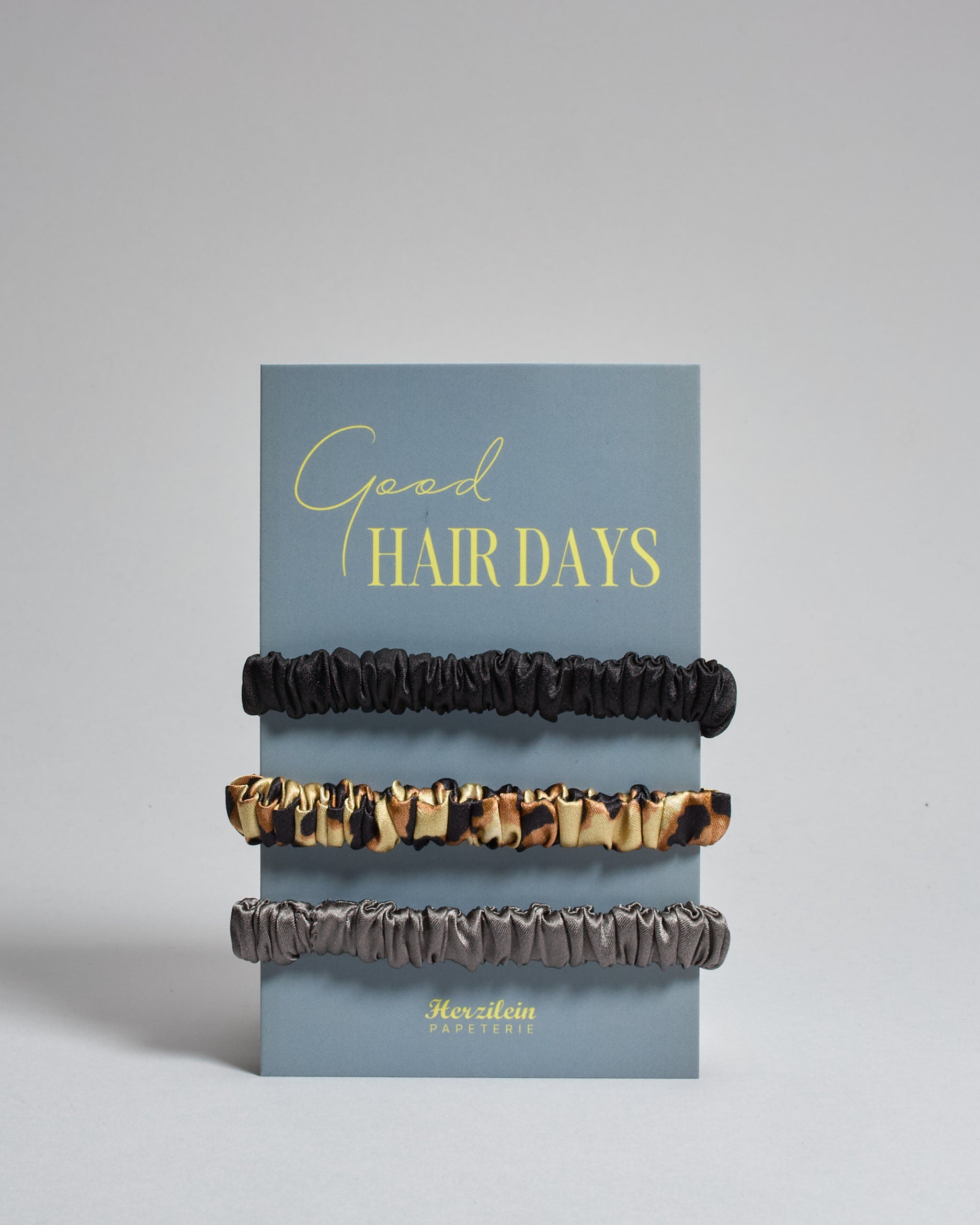 Drei Haar-Scrunchies aus Seide auf einer Karte auf der oben "good hair days" steht. Die Scrunchies sind schwarz, leo und grau. 