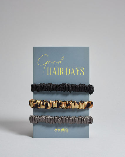 Drei Haar-Scrunchies aus Seide auf einer Karte auf der oben "good hair days" steht. Die Scrunchies sind schwarz, leo und grau. 