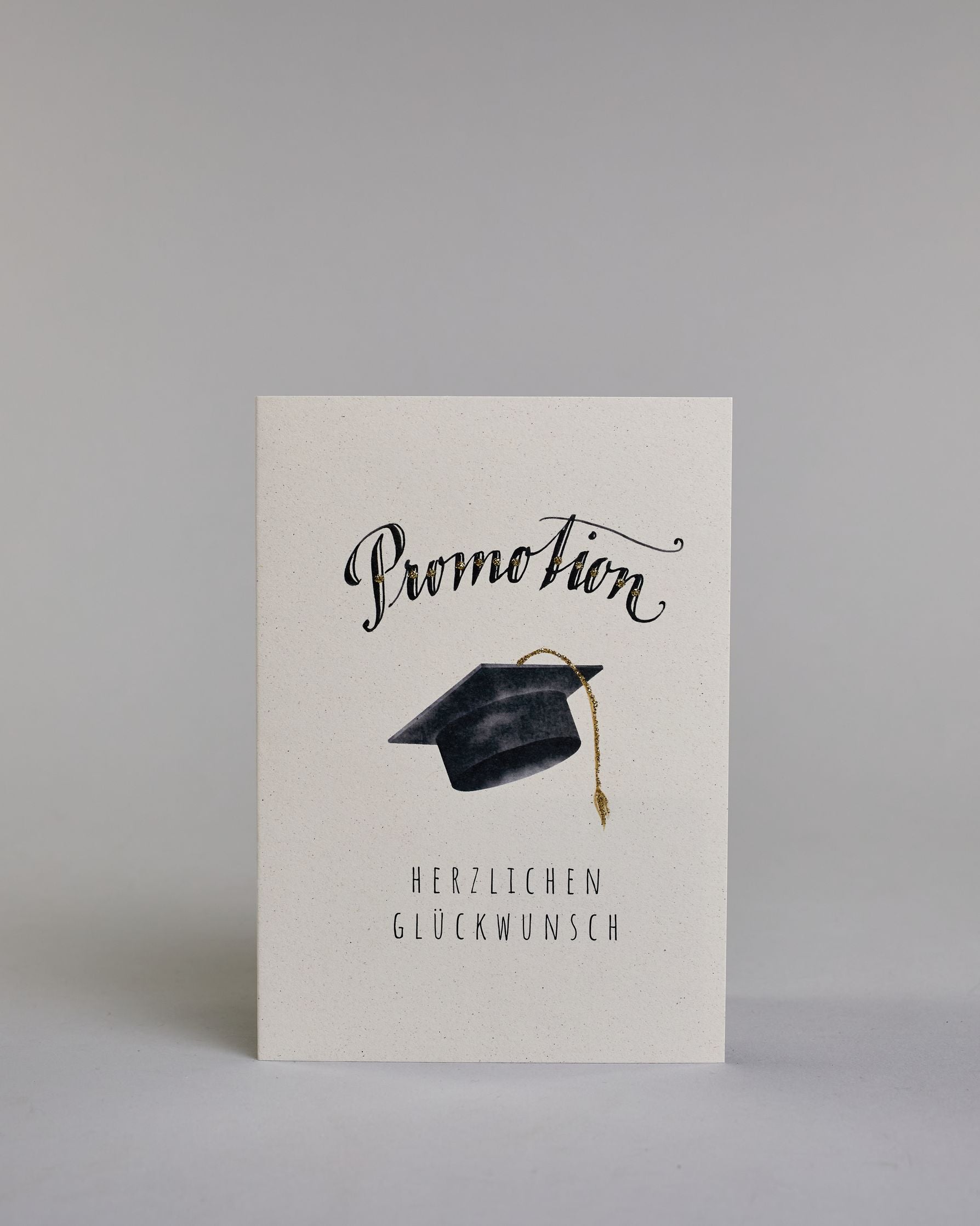 Glückwunschkarte zur Promotion mit schwarzer Kalligrafieaufschrift ‚Promotion‘, darunter ein schwarzer Doktorhut mit goldener Quaste und der Text ‚Herzlichen Glückwunsch‘ auf hellem Hintergrund. Dazu ein grün-goldenes handgeschöpftes Kuvert. #1