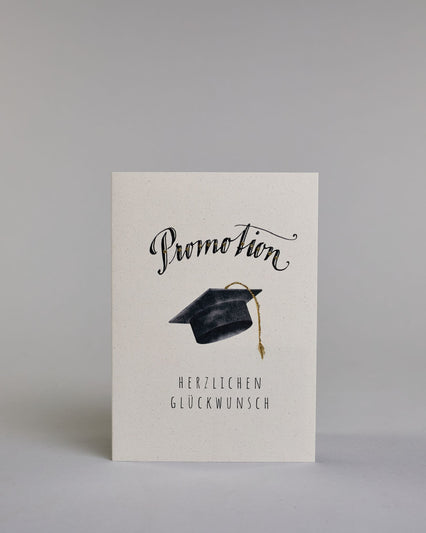 Glückwunschkarte zur Promotion mit schwarzer Kalligrafieaufschrift ‚Promotion‘, darunter ein schwarzer Doktorhut mit goldener Quaste und der Text ‚Herzlichen Glückwunsch‘ auf hellem Hintergrund. Dazu ein grün-goldenes handgeschöpftes Kuvert. #1