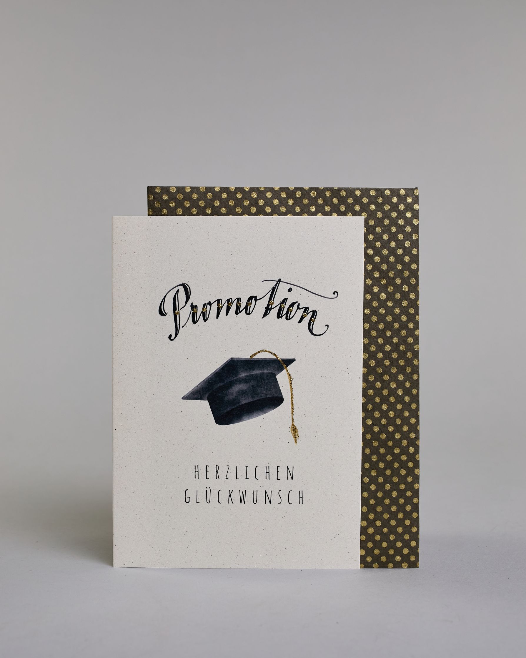 Glückwunschkarte zur Promotion mit schwarzer Kalligrafieaufschrift ‚Promotion‘, darunter ein schwarzer Doktorhut mit goldener Quaste und der Text ‚Herzlichen Glückwunsch‘ auf hellem Hintergrund. Dazu ein grün-goldenes handgeschöpftes Kuvert. #2