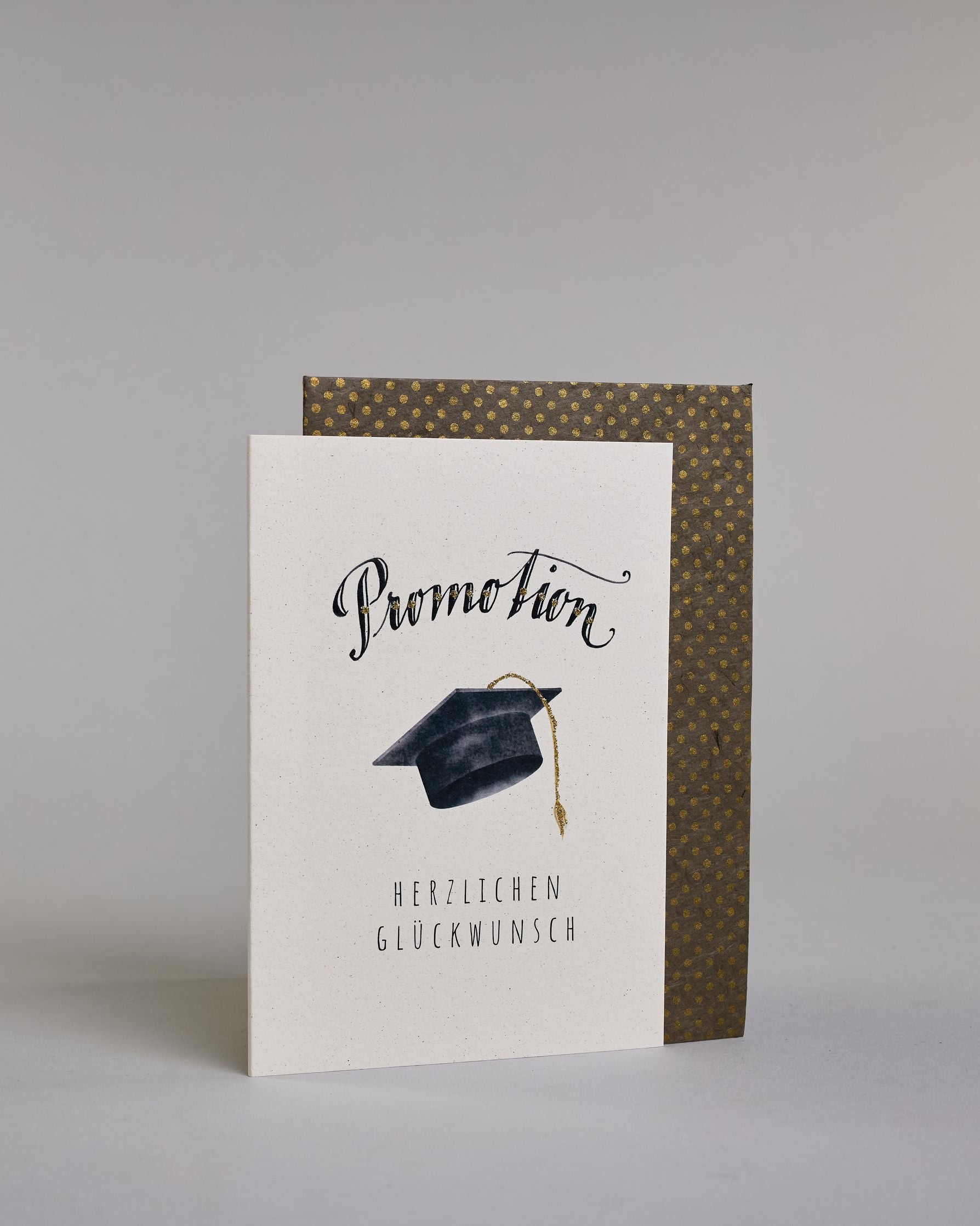 Glückwunschkarte zur Promotion mit schwarzer Kalligrafieaufschrift ‚Promotion‘, darunter ein schwarzer Doktorhut mit goldener Quaste und der Text ‚Herzlichen Glückwunsch‘ auf hellem Hintergrund. Dazu ein grün-goldenes handgeschöpftes Kuvert. #3