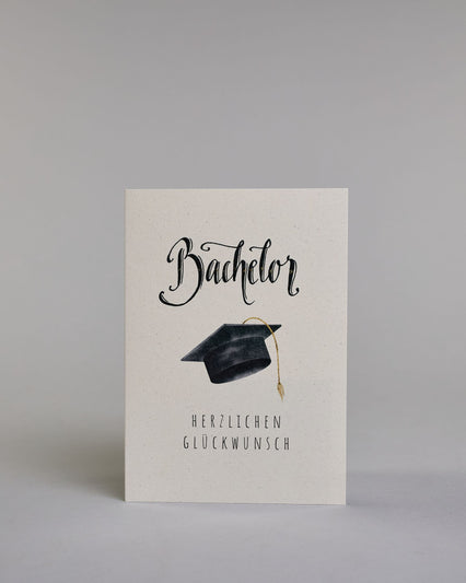 Glückwunschkarte zum Bachelor mit schwarzer Kalligrafieaufschrift ‚Bachelor‘, darunter ein schwarzer Doktorhut mit goldener Quaste und der Text ‚Herzlichen Glückwunsch‘ auf hellem Hintergrund. Dazu ein grün-goldenes handgeschöpftes Kuvert. #1
