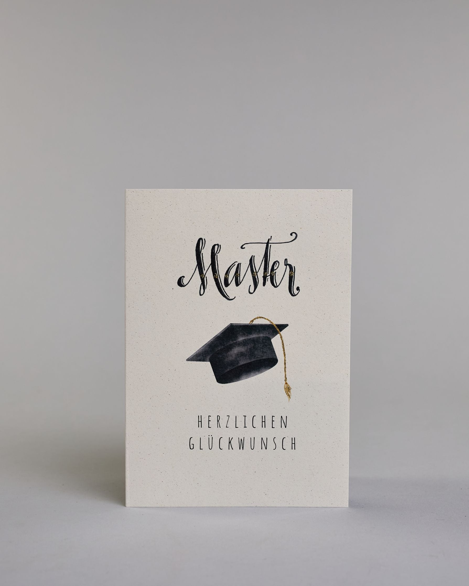Glückwunschkarte zum Master mit schwarzer Kalligrafieaufschrift ‚Master‘, darunter ein schwarzer Doktorhut mit goldener Quaste und der Text ‚Herzlichen Glückwunsch‘ auf hellem Hintergrund. Dazu ein grün-goldenes handgeschöpftes Kuvert. #1