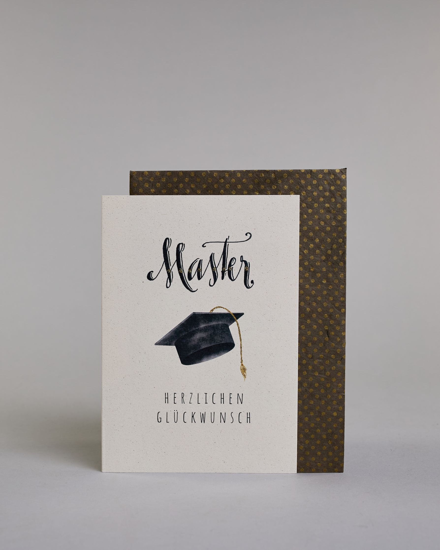 Glückwunschkarte zum Master mit schwarzer Kalligrafieaufschrift ‚Master‘, darunter ein schwarzer Doktorhut mit goldener Quaste und der Text ‚Herzlichen Glückwunsch‘ auf hellem Hintergrund. Dazu ein grün-goldenes handgeschöpftes Kuvert. #2