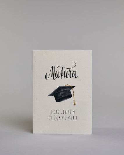 Glückwunschkarte zur Matura mit schwarzer Kalligrafieaufschrift ‚Matura‘, darunter ein schwarzer Doktorhut mit goldener Quaste und der Text ‚Herzlichen Glückwunsch‘ auf hellem Hintergrund. Dazu ein grün-goldenes handgeschöpftes Kuvert. #1