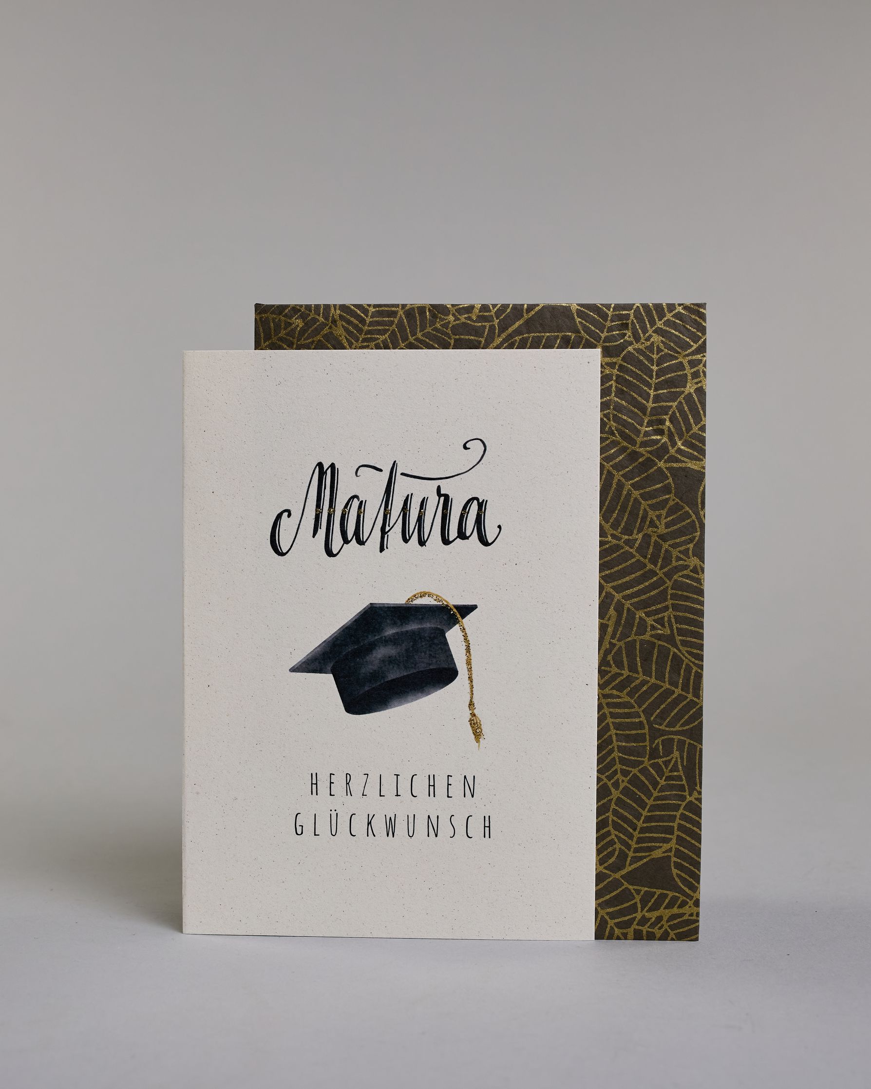 Glückwunschkarte zur Matura mit schwarzer Kalligrafieaufschrift ‚Matura‘, darunter ein schwarzer Doktorhut mit goldener Quaste und der Text ‚Herzlichen Glückwunsch‘ auf hellem Hintergrund. Dazu ein grün-goldenes handgeschöpftes Kuvert. #2